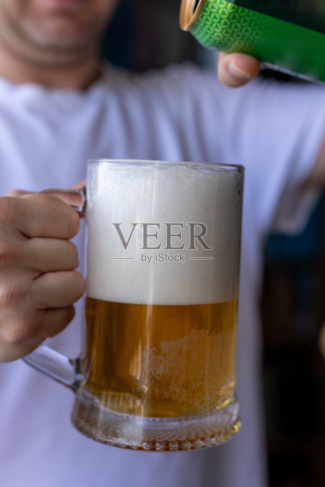 男子将罐装啤酒倒入泡沫丰富的玻璃杯中,前面是一件白色T恤。照片摄影图片