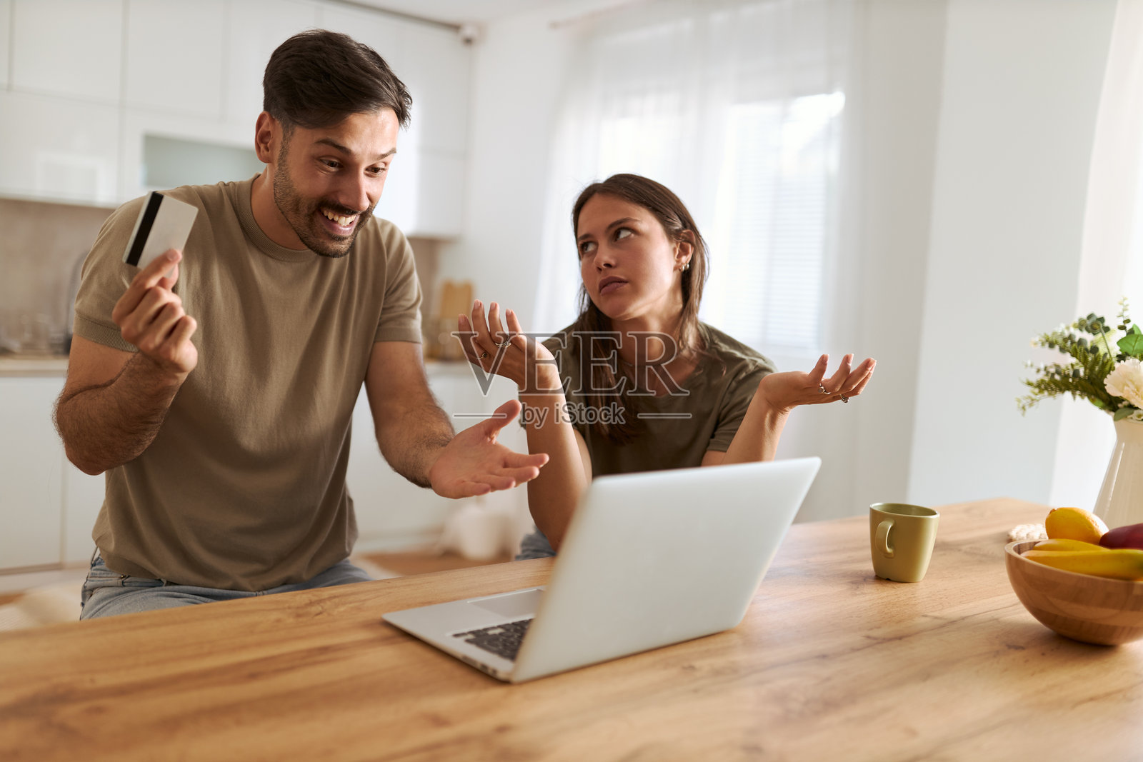 微笑的男人在家中使用信用卡通过笔记本电脑进行在线购物,而他的妻子显得不满。照片摄影图片