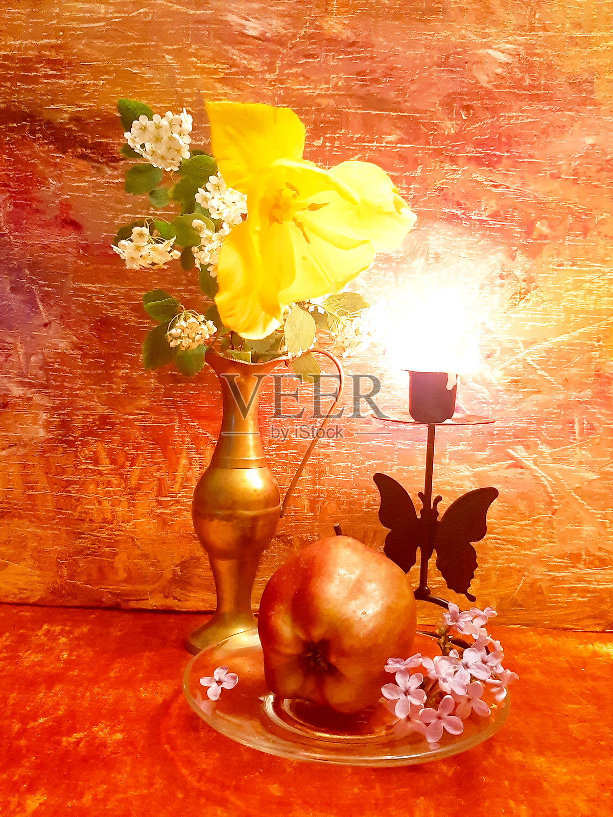一束花、一只苹果、一根燃烧的蜡烛在烛台中。明亮浪漫的静物画,红色背景上的蜡烛与烛台。浪漫、花卉、优雅、风格。古董黄铜。照片摄影图片