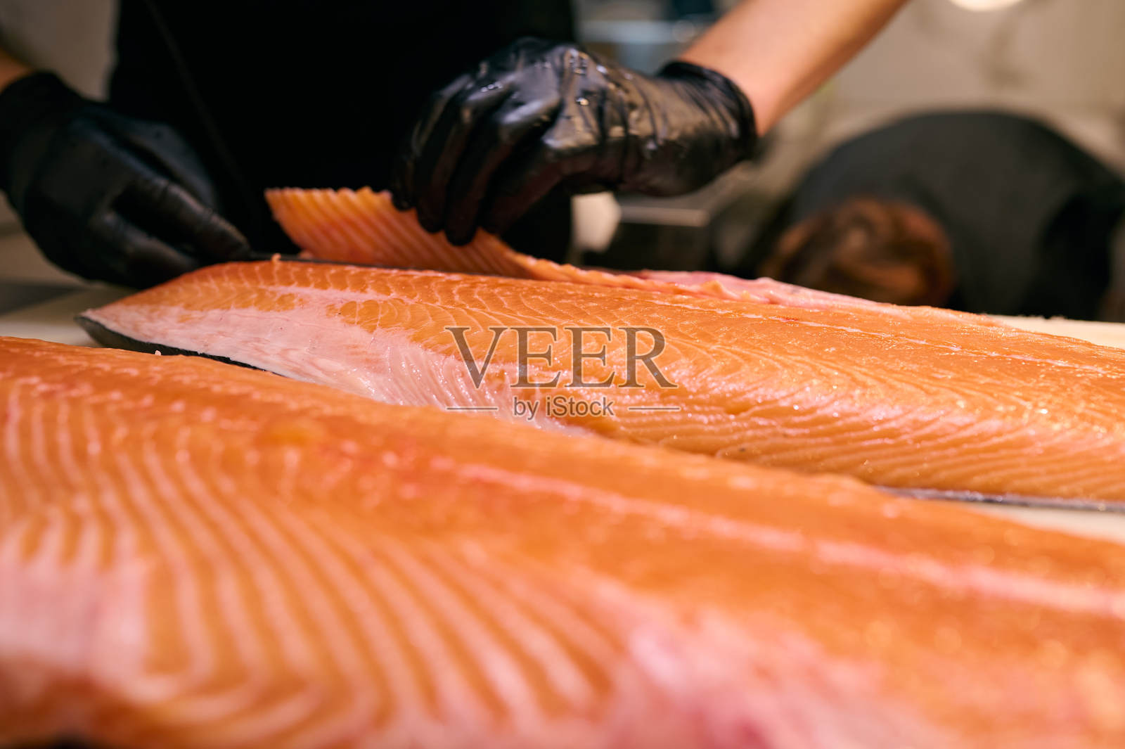 厨师在专业厨房的切菜板上精准地切割新鲜的三文鱼片。特写镜头展示了戴着黑色手套的手和生鱼的纹理。照片摄影图片