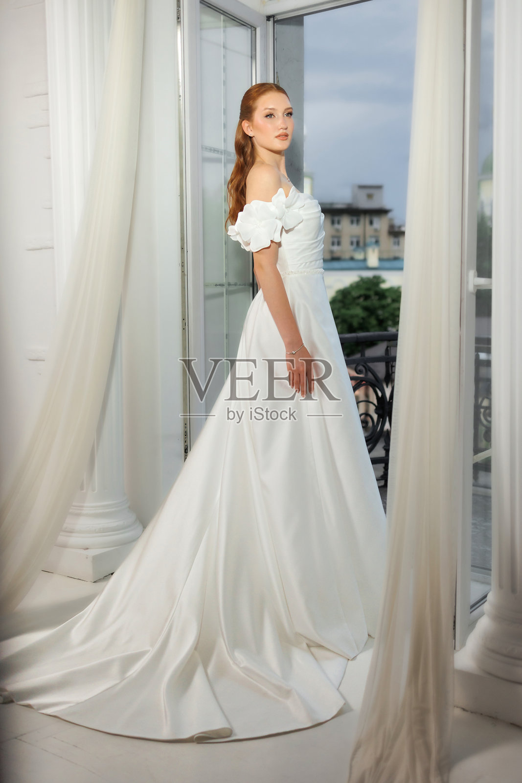 优雅的新娘身着飘逸的白色婚纱,在黄昏时分的开放窗边优雅地摆姿势,窗外是城市的美景。照片摄影图片