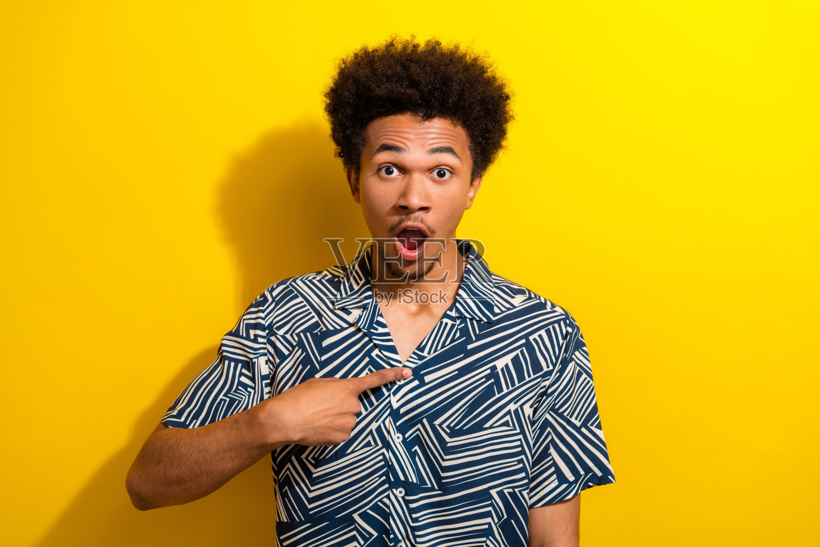 穿着图案衬衫的年轻男子惊讶地指着自己,背景是鲜艳的黄色。照片摄影图片