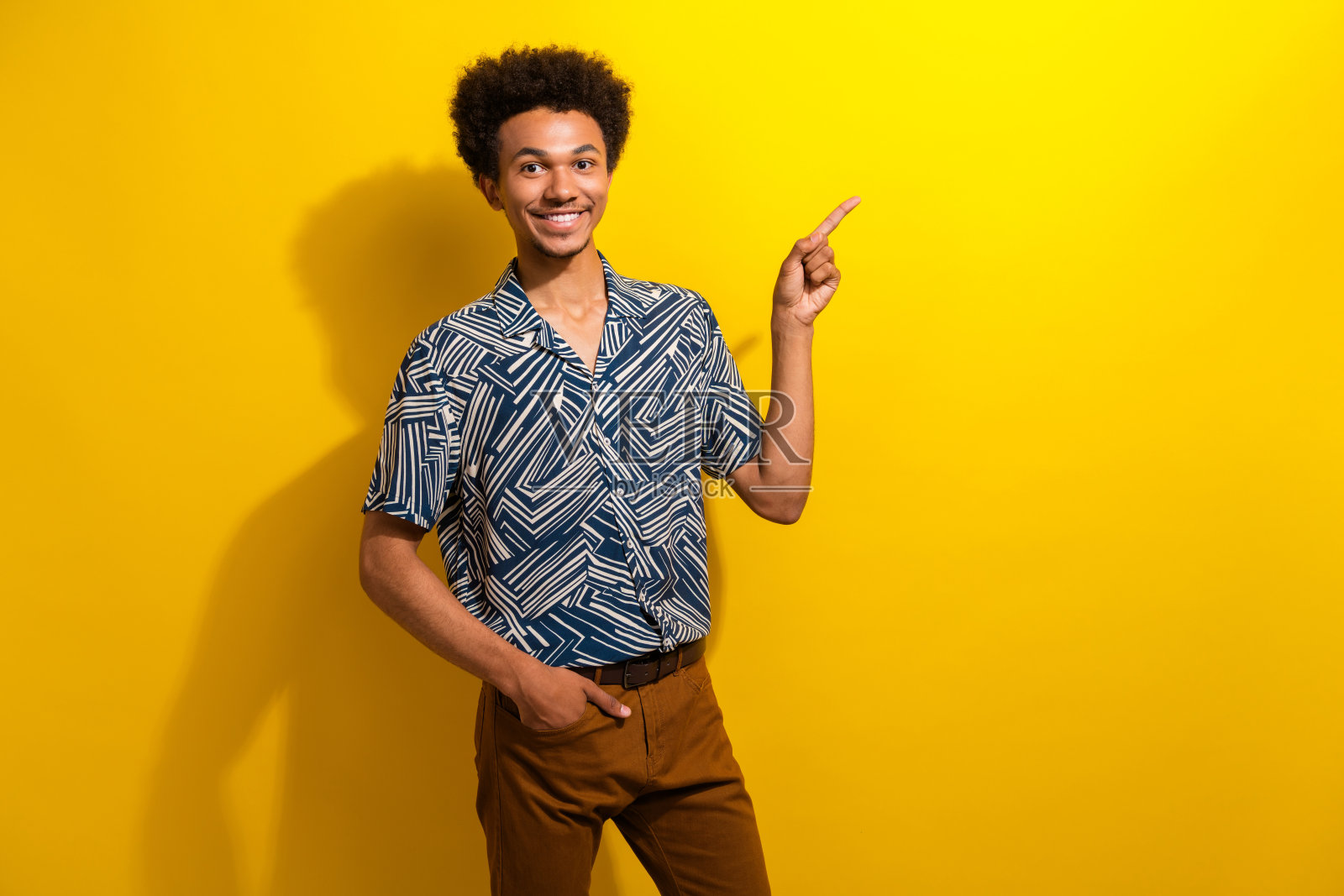 充满魅力的年轻男子穿着时尚的服装,面带愉快的表情,指向一个方向,背景是明亮的黄色。照片摄影图片