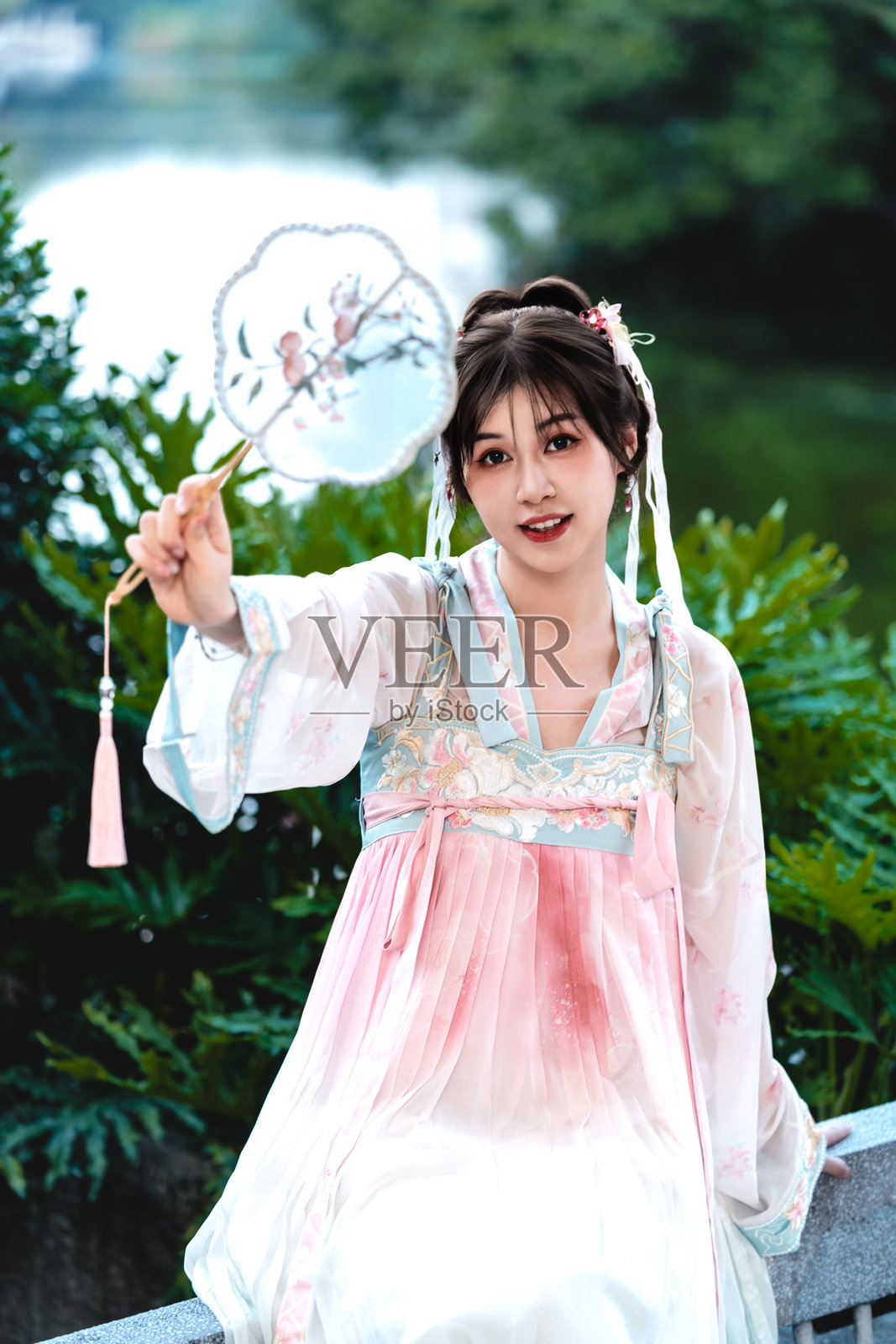 身穿经典唐汉服的美丽亚洲女性,手持扇子,坐在传统中国园林中的石凳上。照片摄影图片