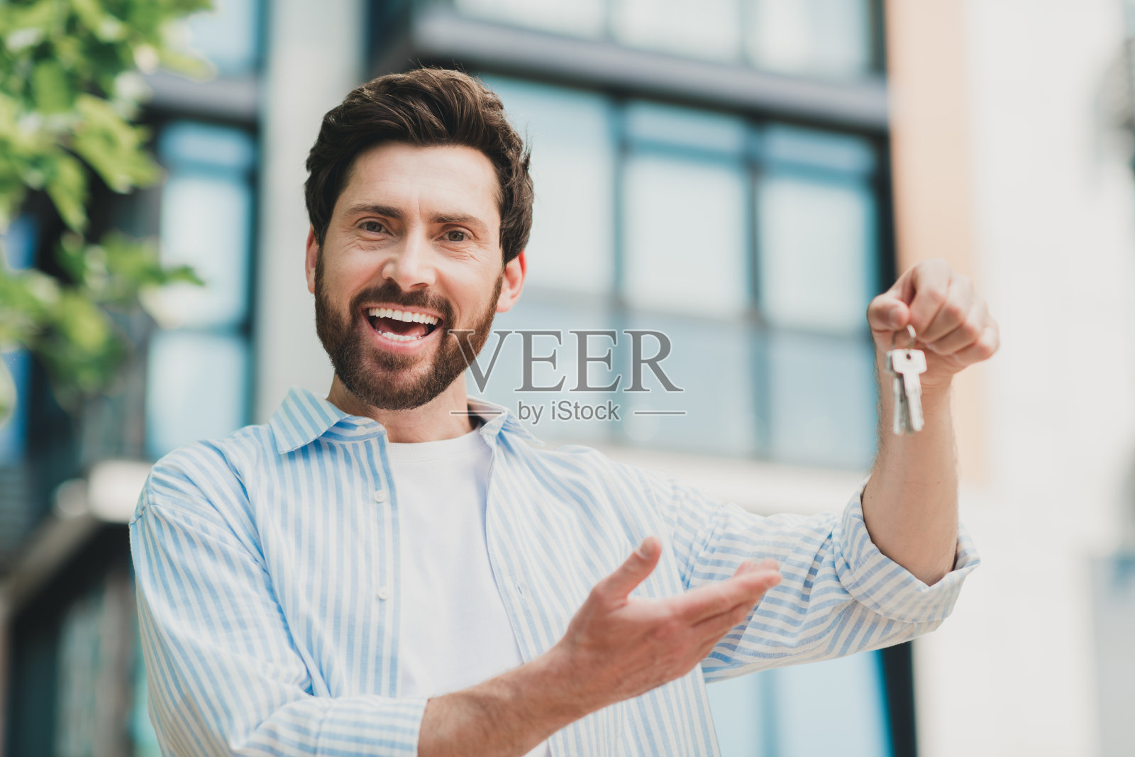 年轻男子在户外自信地微笑,手持钥匙并朝阳光明媚的城市建筑做出手势。照片摄影图片