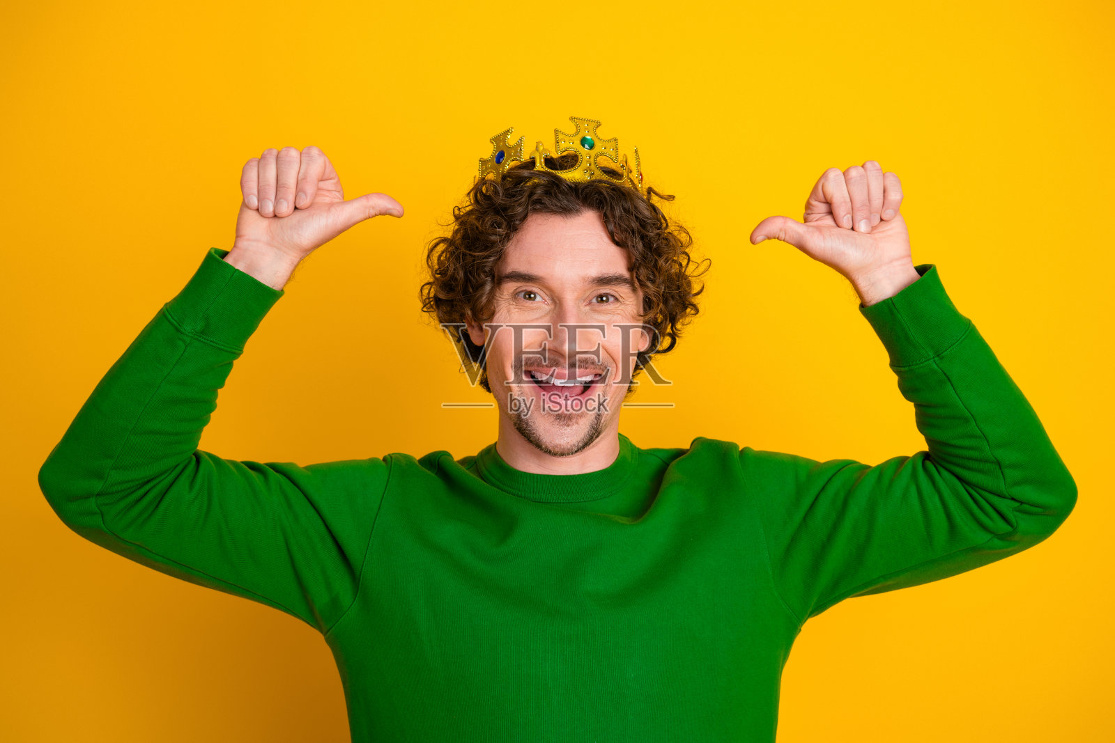 穿着绿色毛衣的快乐年轻男子,头戴王冠,调皮地对自己竖起大拇指,背景为黄色。照片摄影图片
