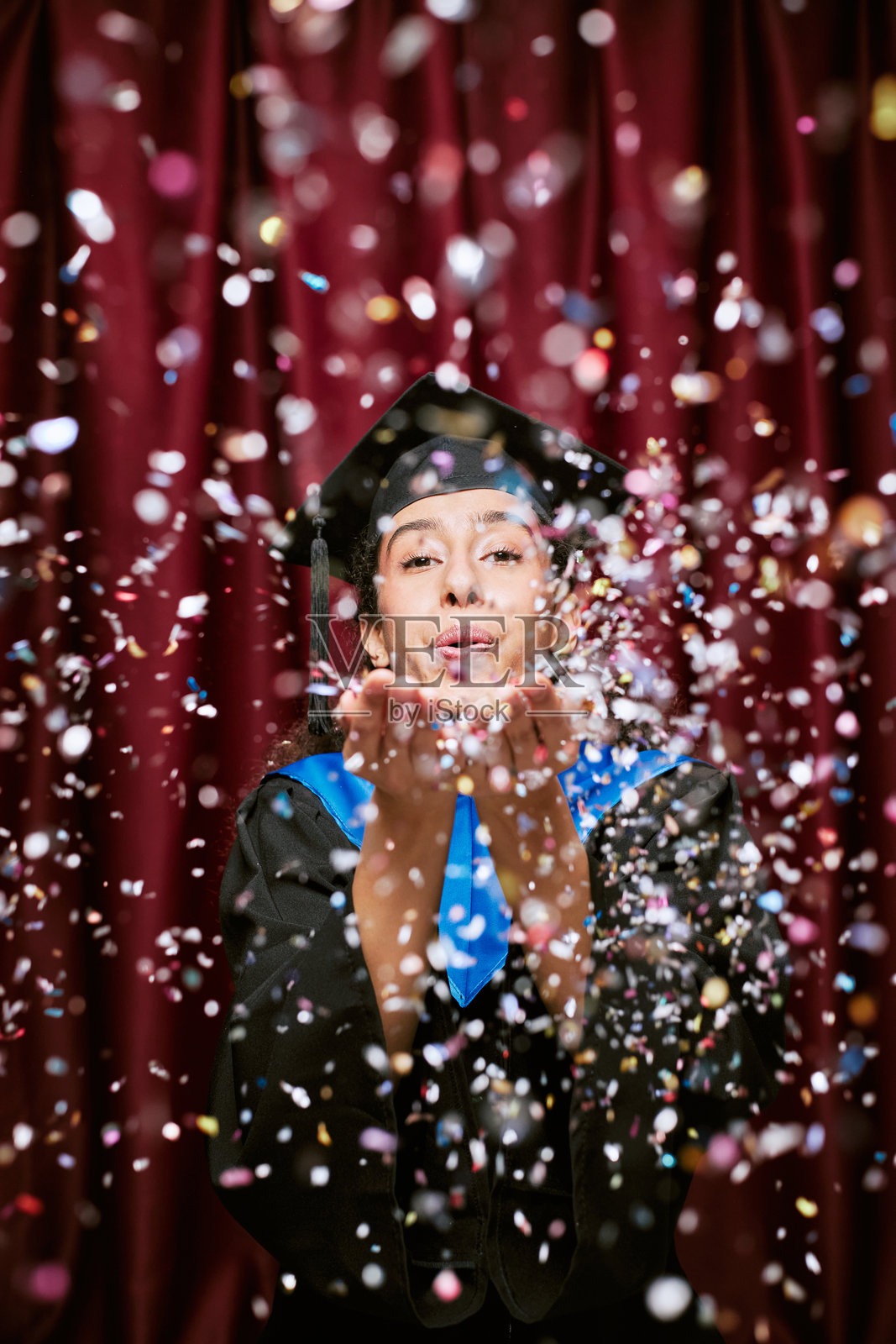 年轻的西班牙裔女性在照片亭庆祝毕业,吹着五彩纸屑照片摄影图片