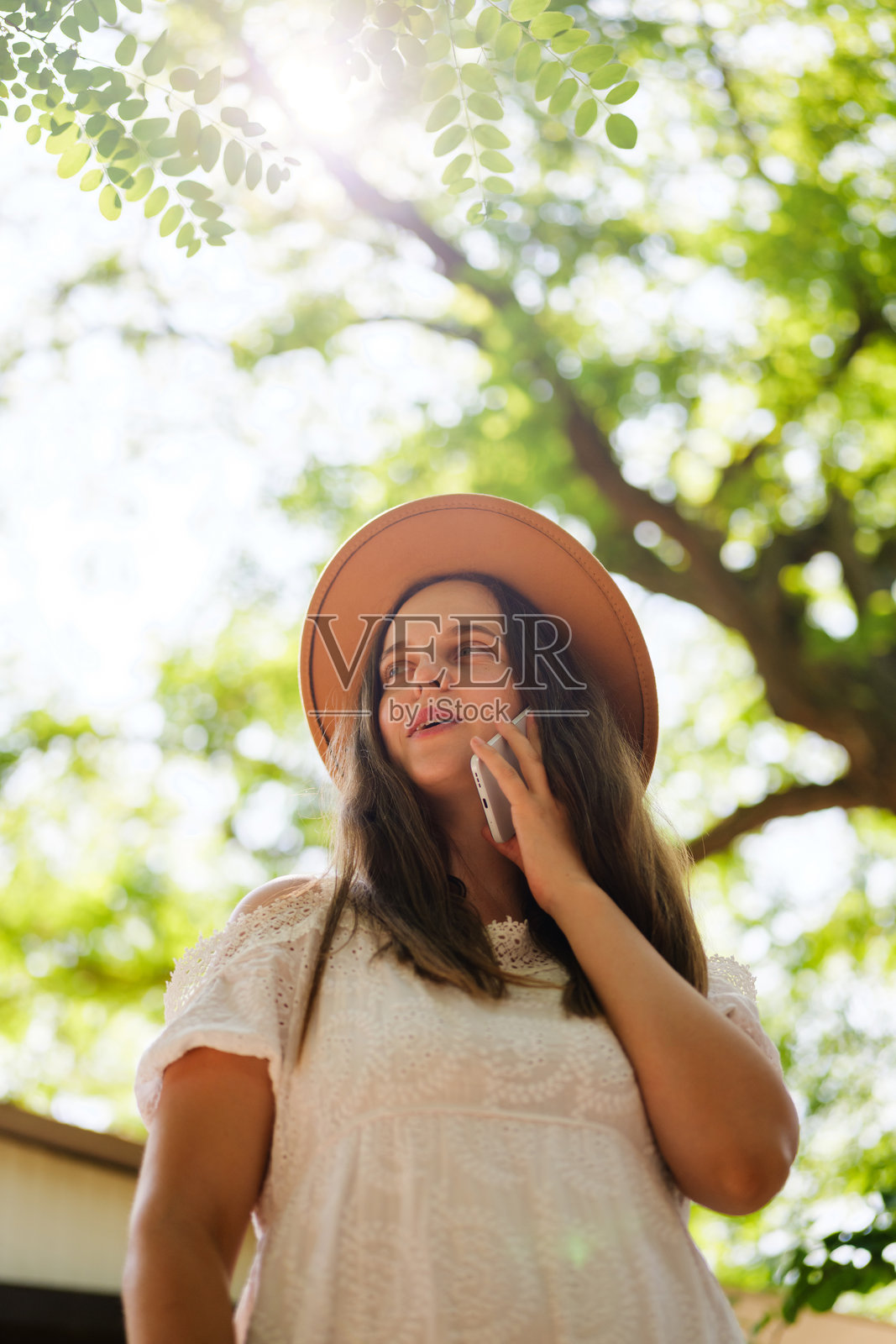 一位穿着白色连衣裙和草帽的年轻女性在阳光斑驳的树叶间,若有所思地看着天空,正在用智能手机通话。照片摄影图片