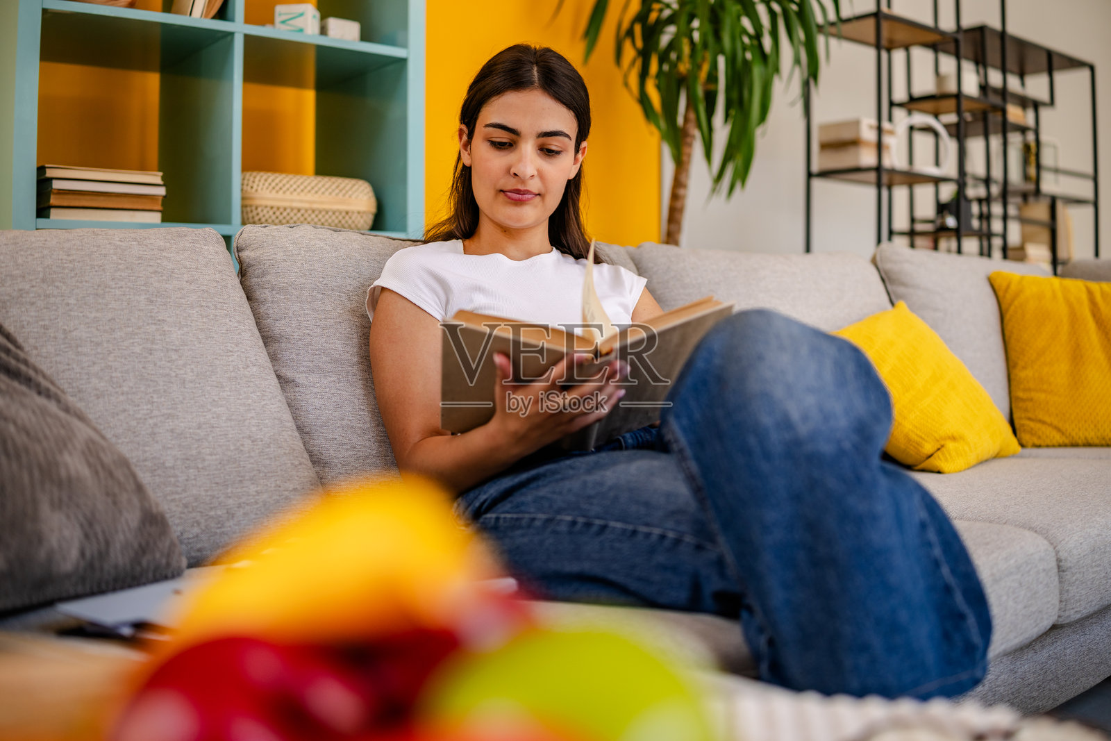 年轻女性在一个充满生机色彩和愉快装饰的舒适客厅里享受阅读,悠闲地坐在舒适的沙发上。照片摄影图片
