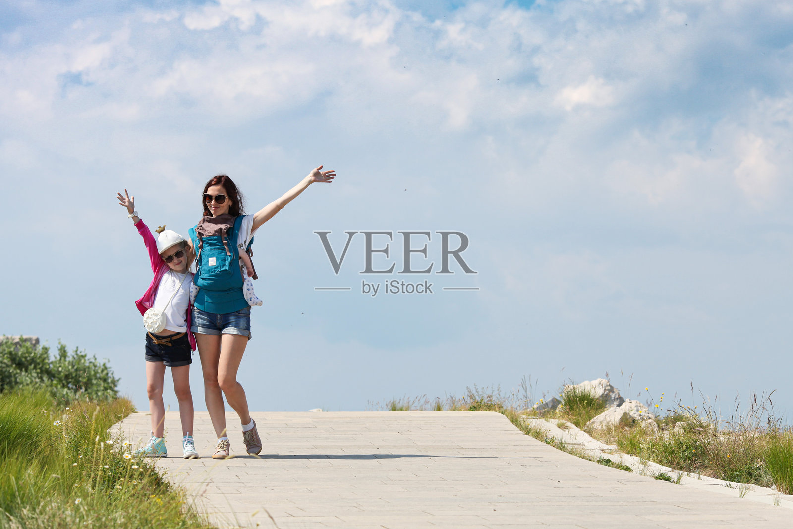 年轻女性背着婴儿,女儿在山顶观景台上举起手臂拍照。照片摄影图片