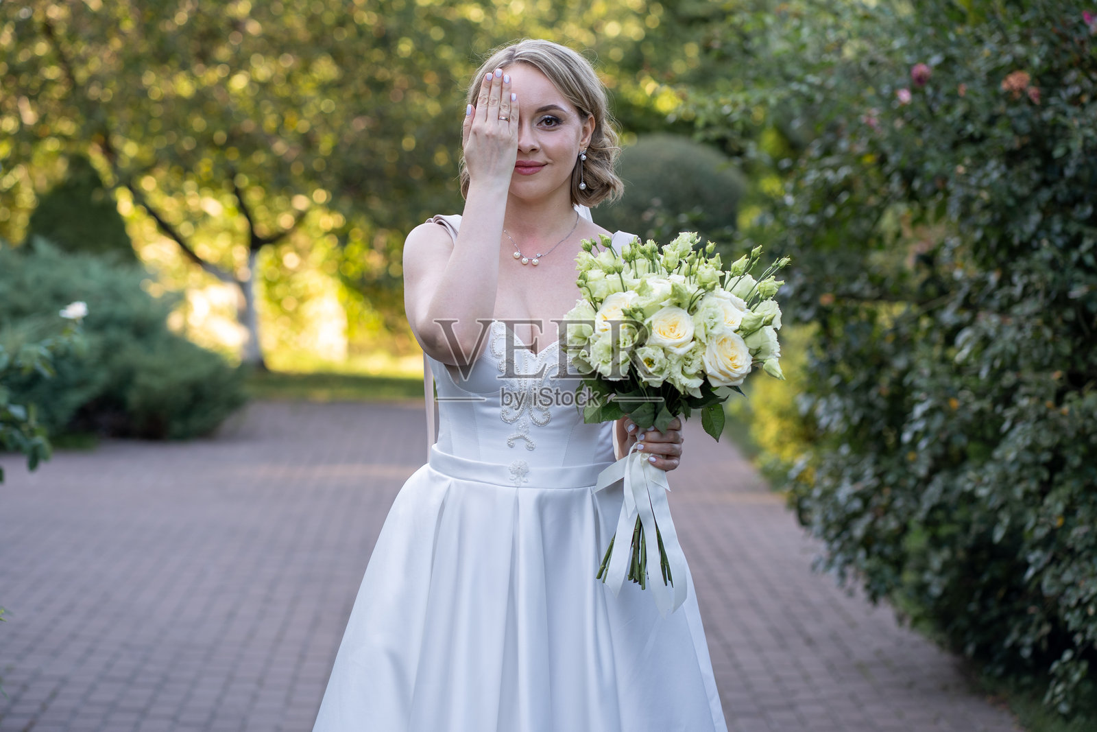 穿着白色茶长婚纱的俏皮新娘,手捧象牙色和淡粉色玫瑰花束,微笑着用手遮住一只眼睛,展示她的婚戒。照片摄影图片