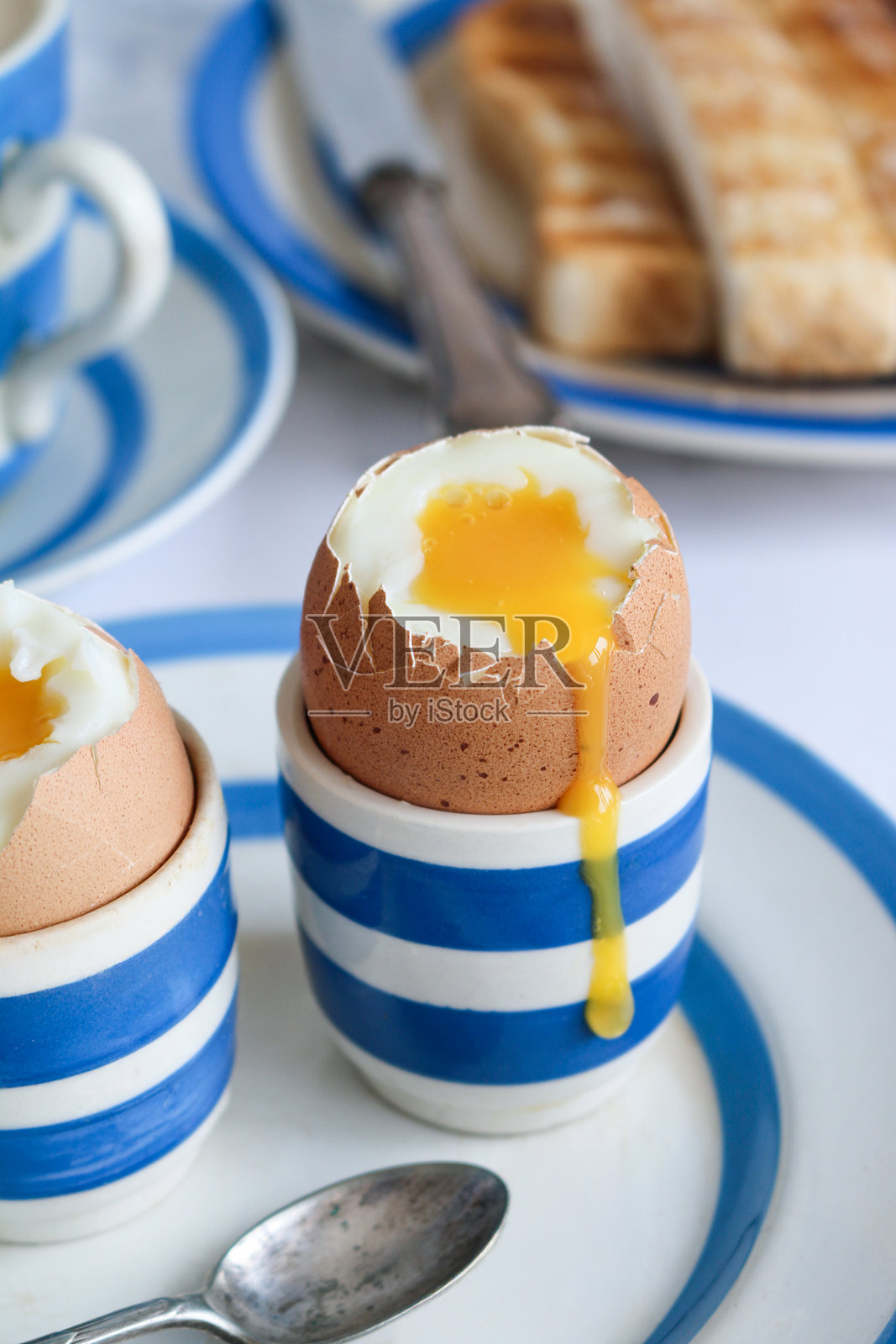 特写镜头展示蓝白色的康沃尔陶器,茶盘和盛有流心煮蛋的蛋杯,茶匙,涂了黄油的白色吐司士兵,刀,盛有奶茶的茶杯和碟子,背景为白色大理石效果,重点在前景。照片摄影图片