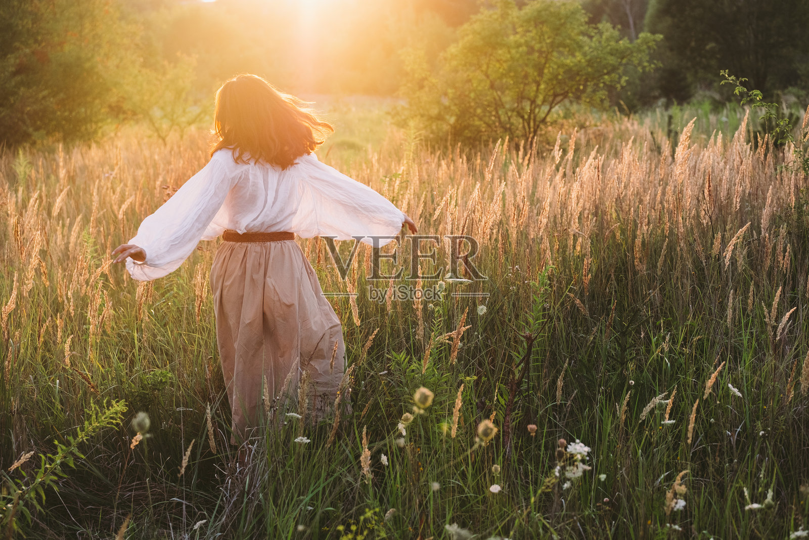 年轻女性背影穿着宽松的波西米亚风格上衣和米色裙子,自由地走在草地上,沐浴在温暖的夕阳下,展现出自由、宁静、正念与自然和谐的氛围。照片摄影图片