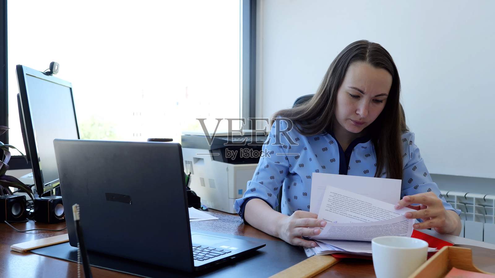 专注的女商人在现代办公室桌前工作,检查文件,旁边有电脑、打印机和网络摄像头。照片摄影图片