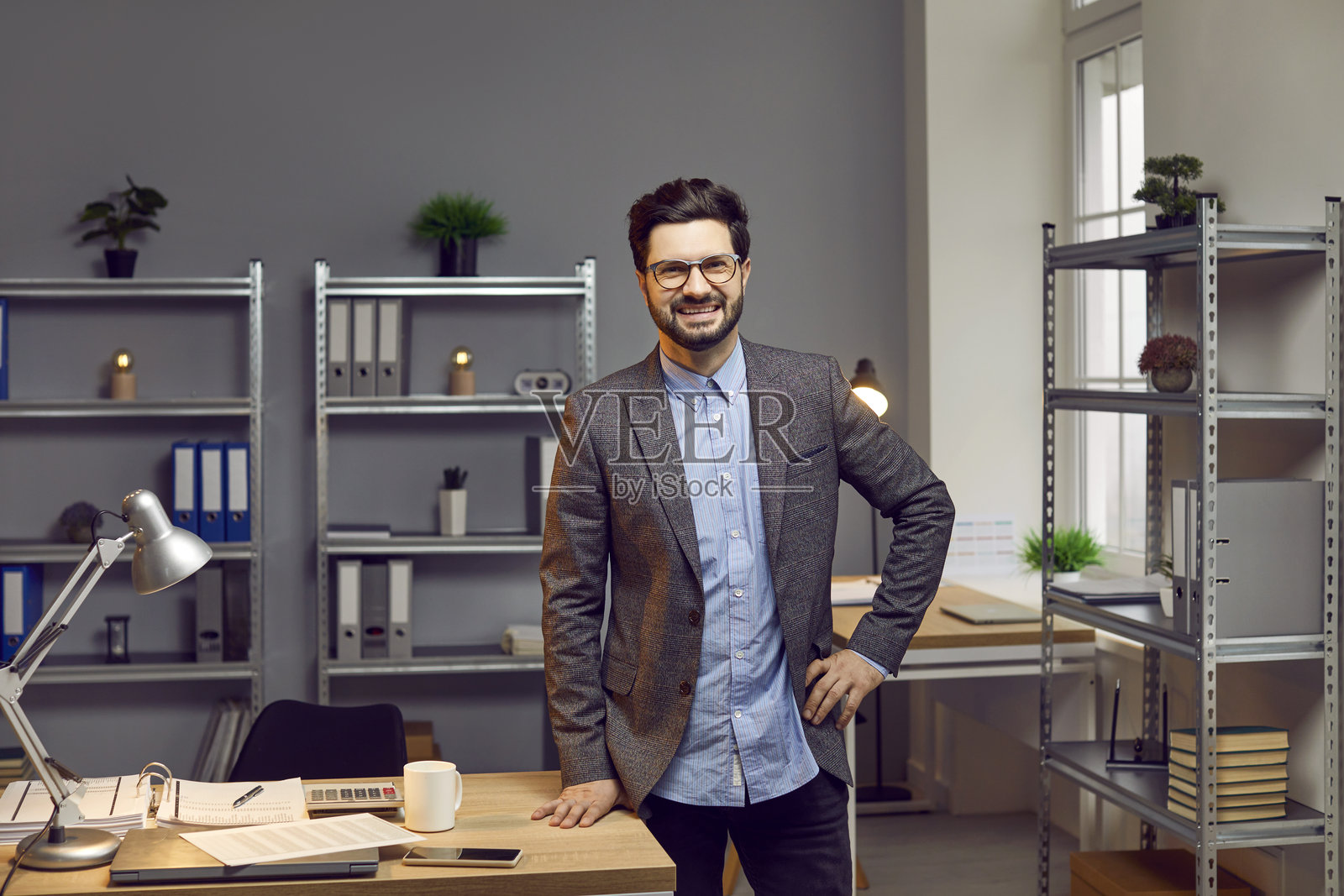 微笑、自信的年轻商人戴着眼镜,靠在宽敞创意办公室的桌子上,桌上摆放着文具。照片摄影图片