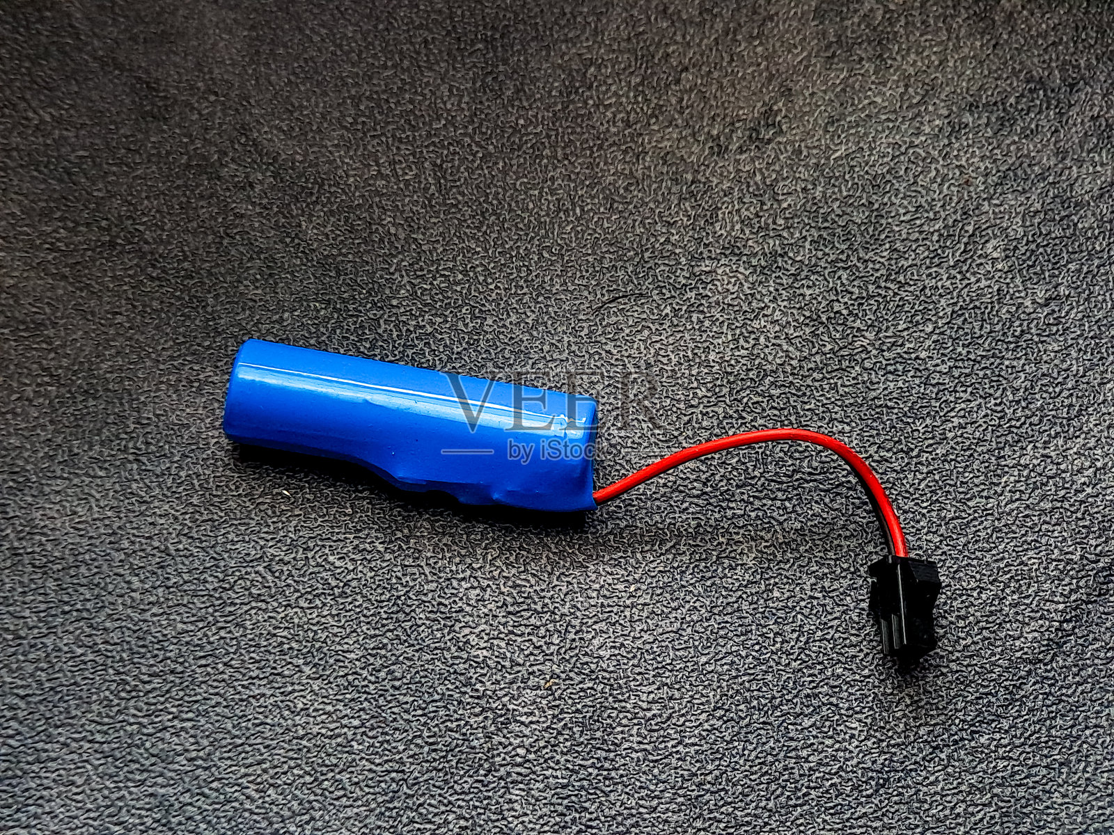 生动的圆柱形电池,外壳为明亮的蓝色,通过一根光滑的红色电线连接到一个小黑色连接器,静静地放在一个有纹理的深色表面上,展示出其功能设计。照片摄影图片