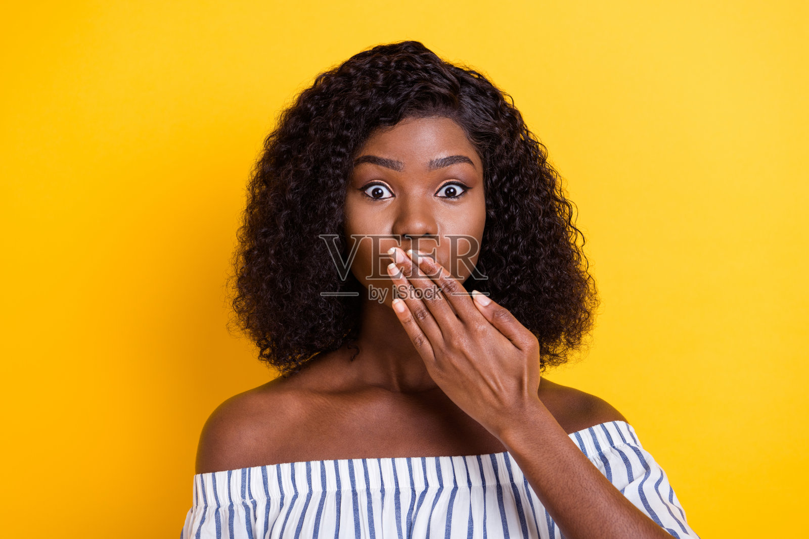 一位肤色较深的迷人女士用手臂遮住嘴唇,神情忧虑地看向镜头,背景为黄色。照片摄影图片