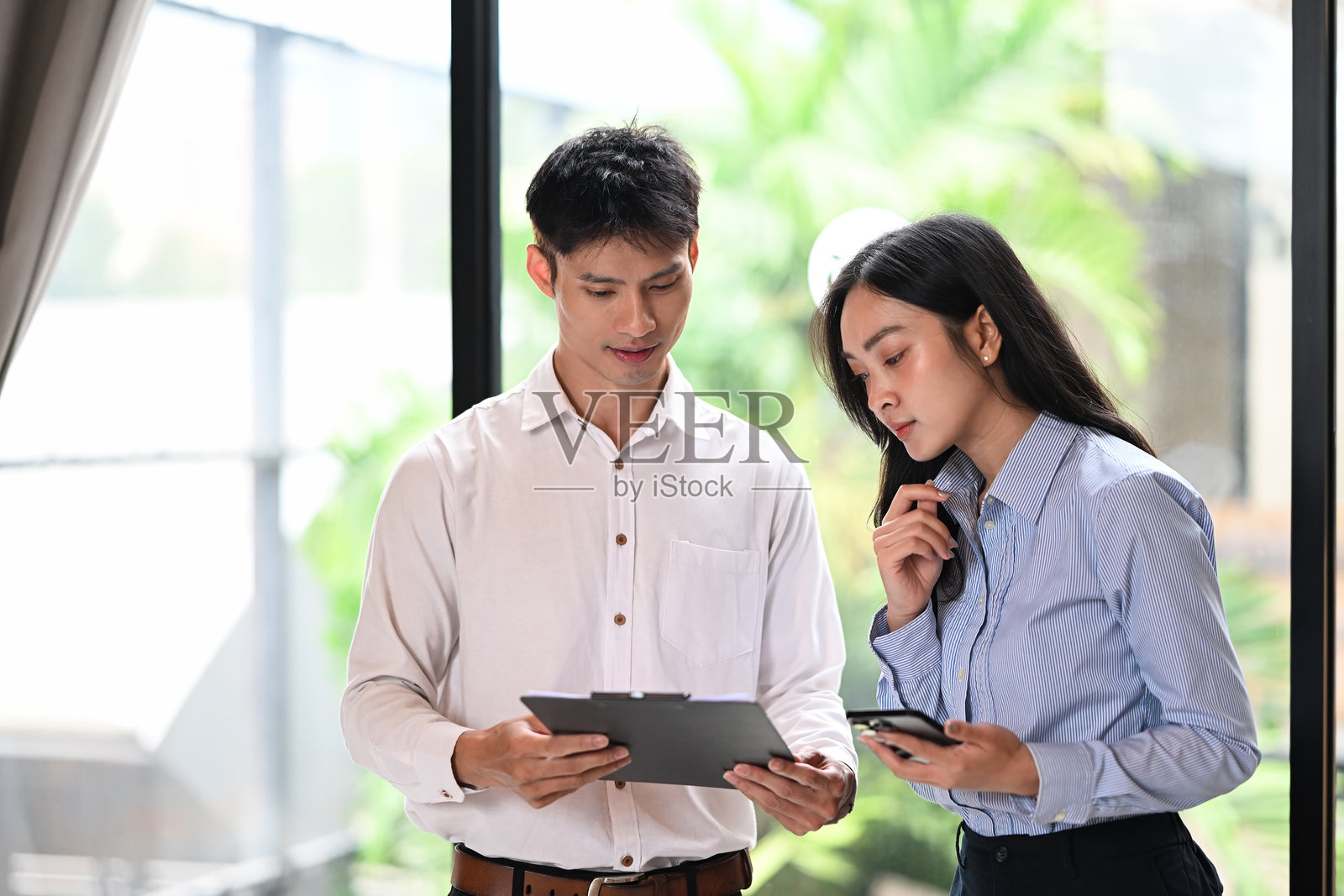 亚洲同事在轻松的工作讨论中查看报告,同时使用手机。照片摄影图片