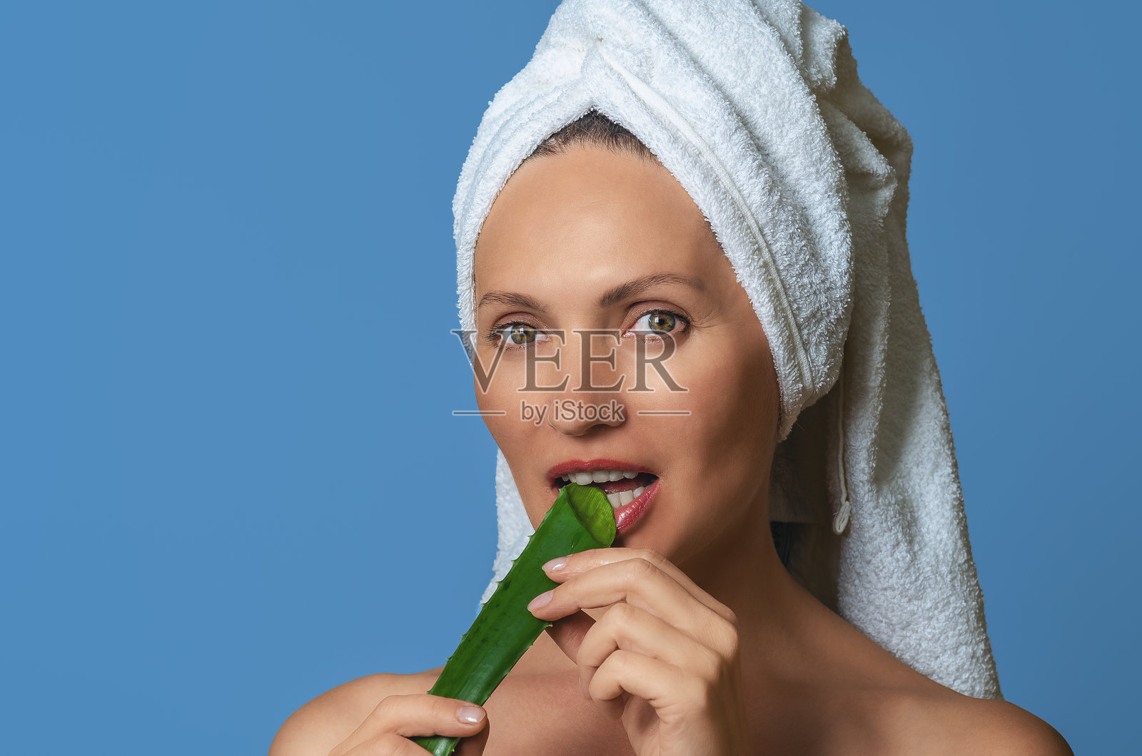 微笑的女性肖像,咬着芦荟叶,洁白的牙齿映衬在蓝色背景上。抗衰老护理概念。照片摄影图片