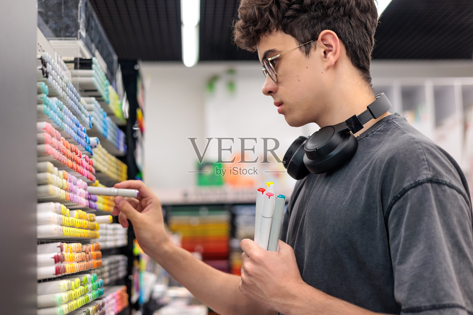 戴着耳机和眼镜的少年站在艺术用品店,挑选五颜六色的马克笔。创造力、爱好、设计、教育和艺术生活方式的概念。照片摄影图片