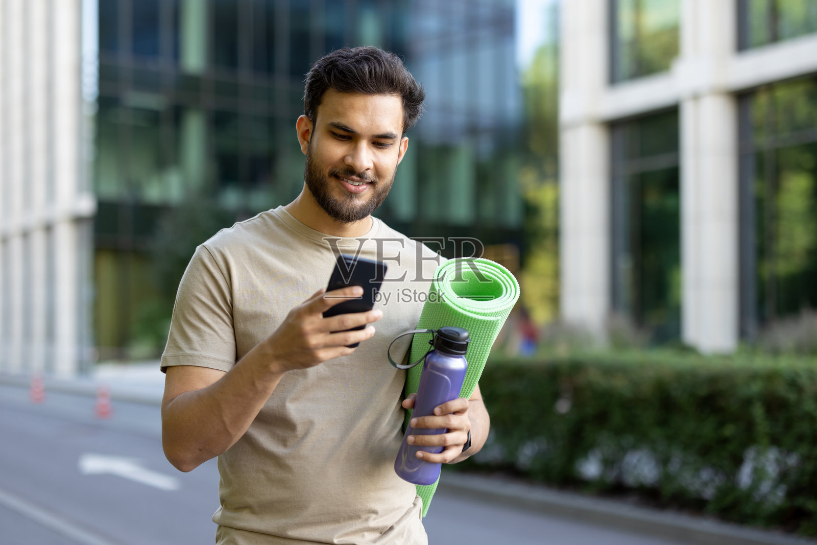 印度年轻男子站在街道上,手里拿着瑜伽垫和水瓶,同时使用手机。照片摄影图片