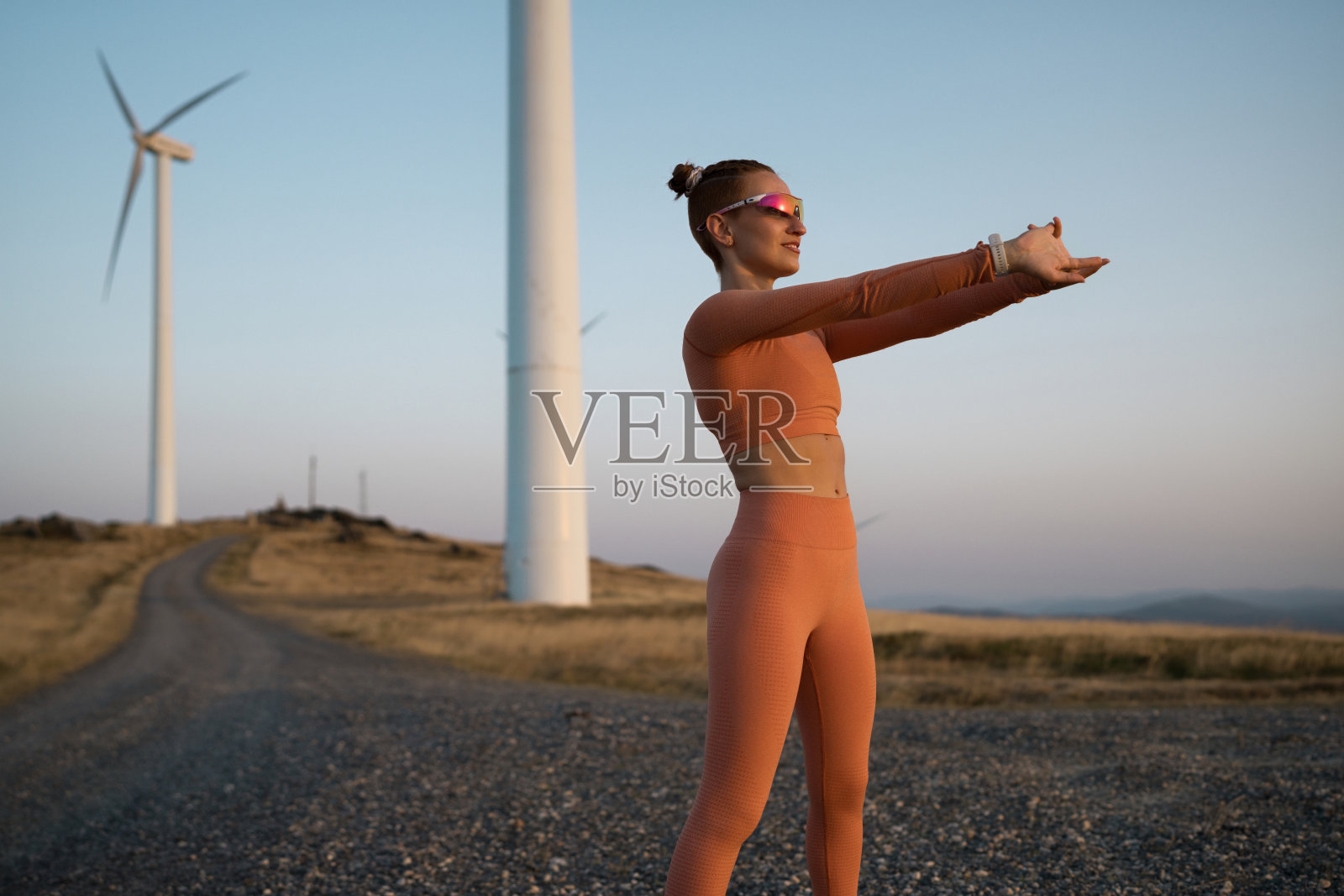 年轻女运动员在日落时分靠近风力发电机拉伸身体照片摄影图片