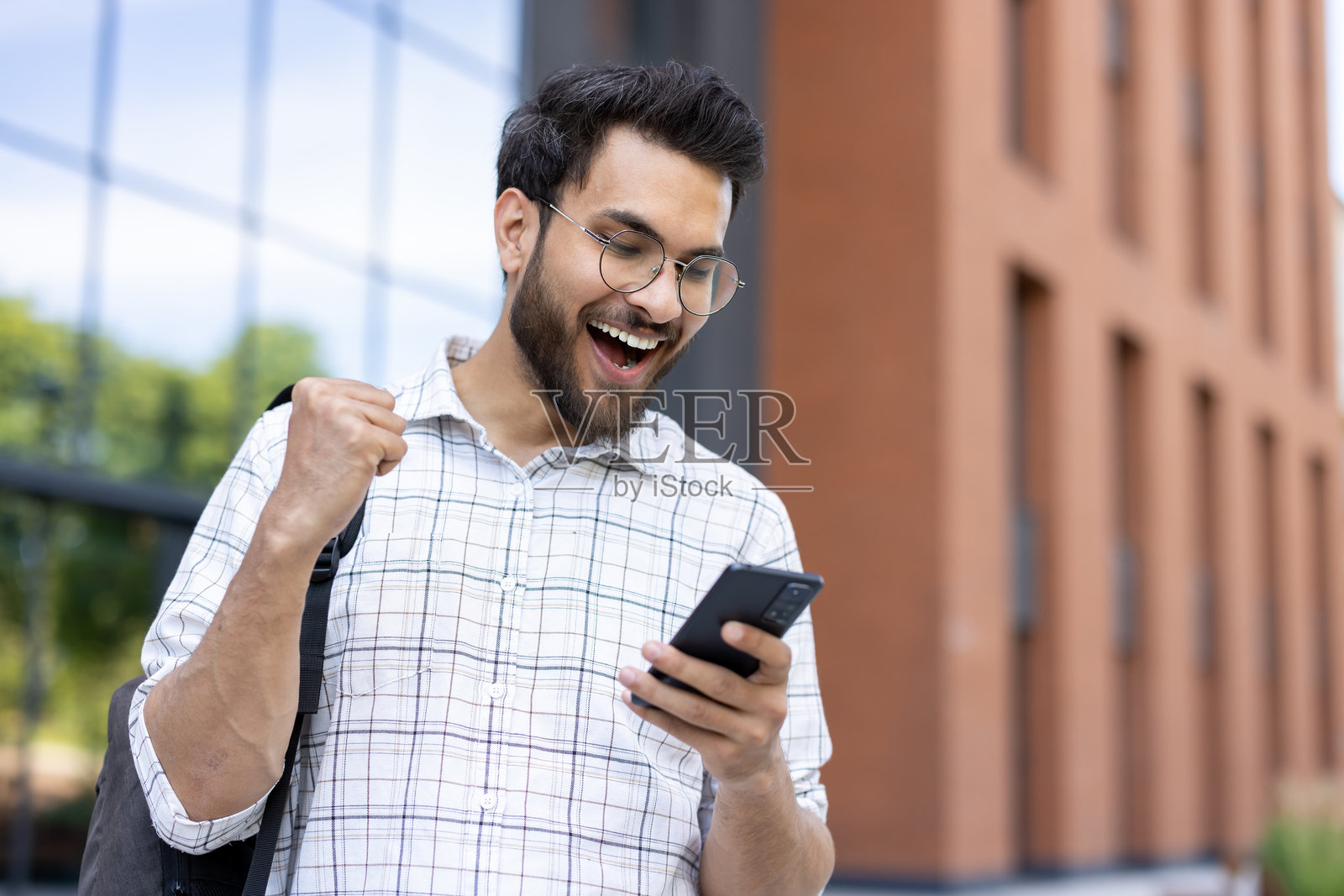 一名年轻学生在建筑外阅读手机时欣喜若狂,庆祝胜利。照片摄影图片