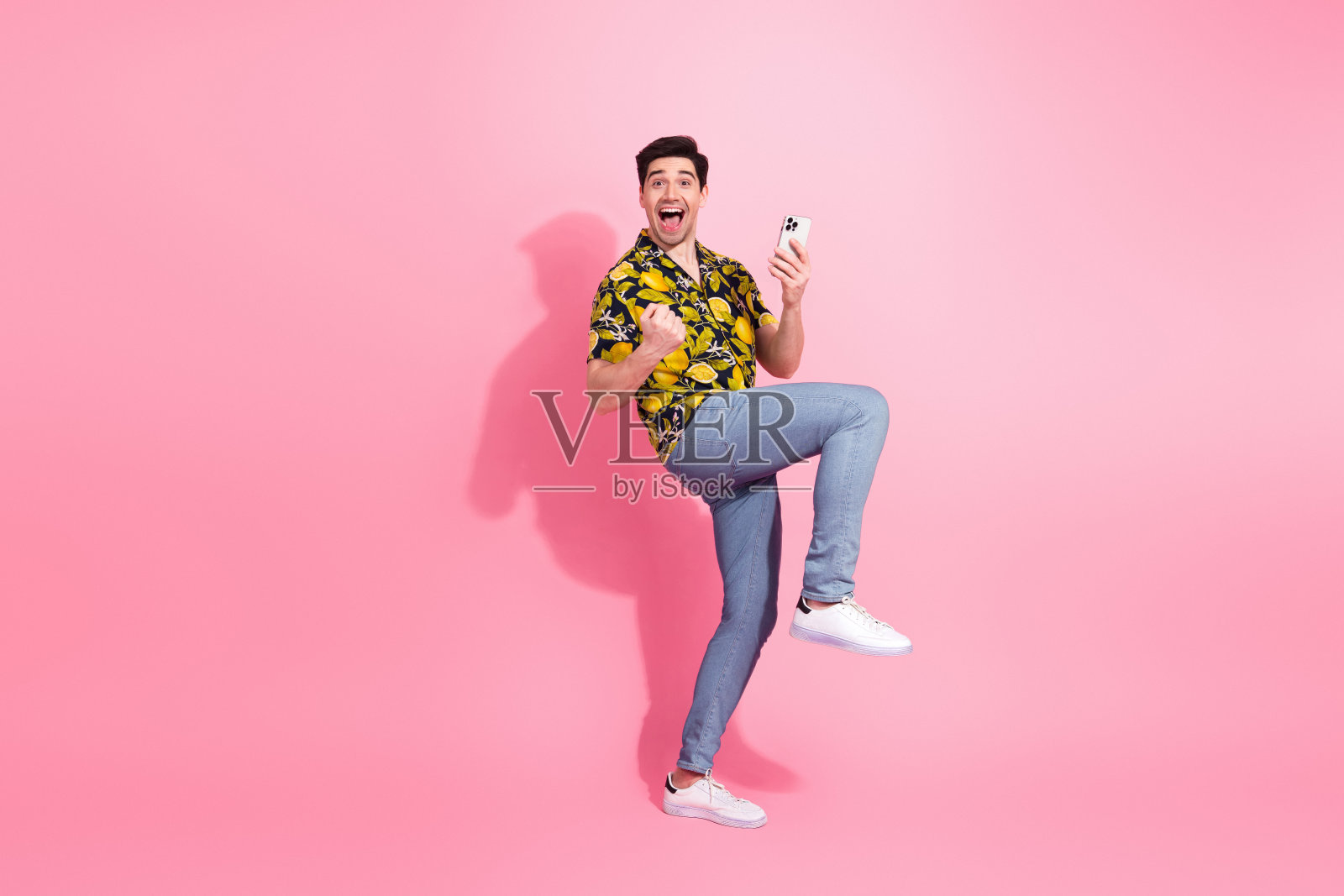 年轻快乐的男士在粉色背景前摆姿势,手持智能手机,展现出时尚的服装和愉悦的心情。照片摄影图片
