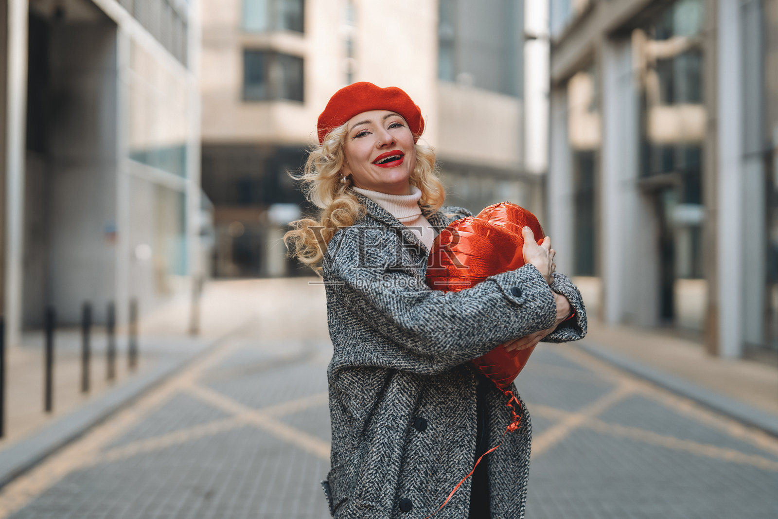 身穿时尚外套和红色贝雷帽的女性在阳光明媚的城市环境中快乐地举着红色心形气球照片摄影图片
