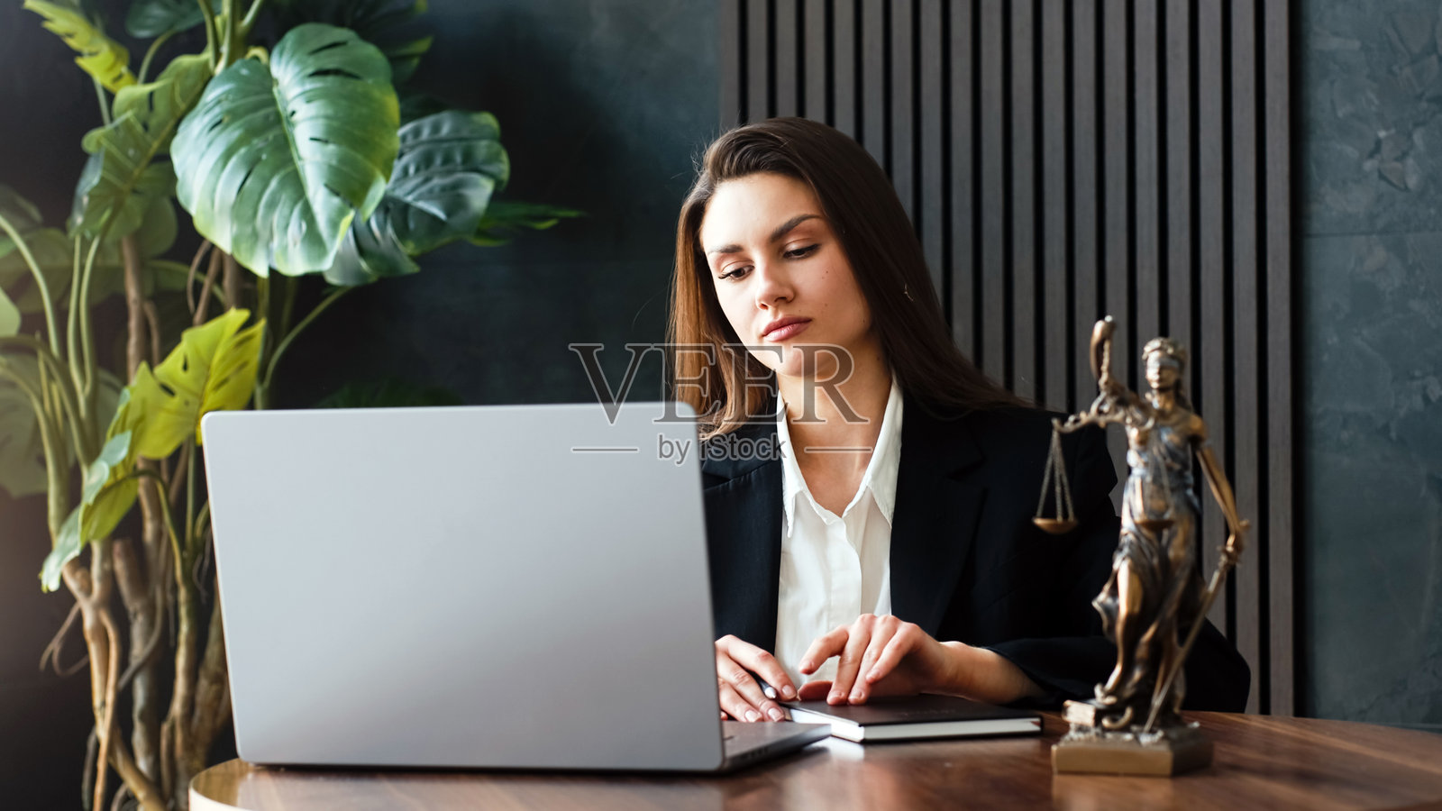 身穿黑色西装的女性律师专注于桌上的笔记本电脑,桌上有法律雕像和绿色植物。照片摄影图片