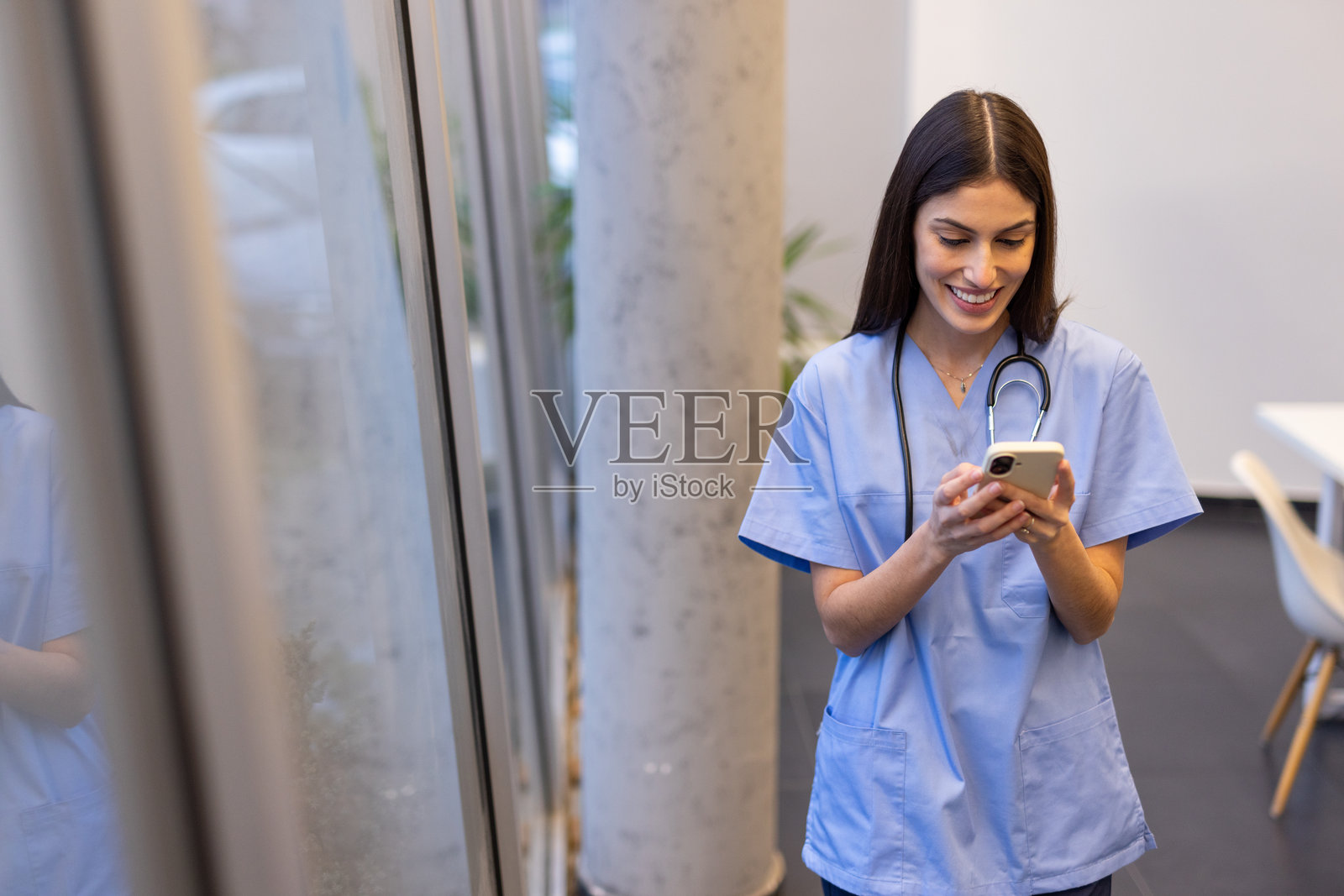 穿着蓝色医务制服的女性医生,在明亮的医疗设施走廊中走动时查看智能手机照片摄影图片