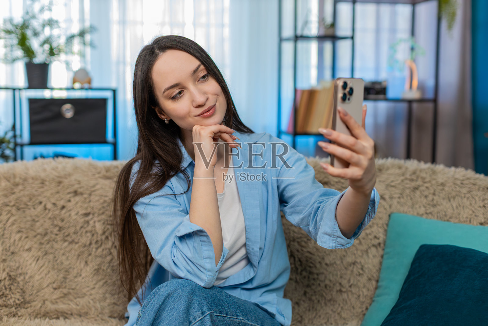 年轻漂亮的女人坐在沙发上,通过视频通话调皮地眨眼微笑,优雅地整理头发。照片摄影图片