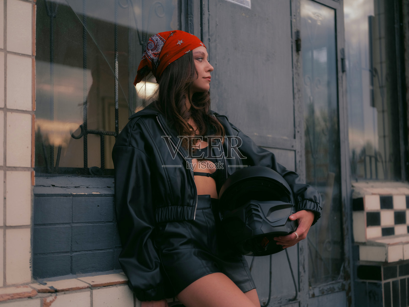 时尚年轻女性穿着皮革服装,头戴红色头巾,户外手持摩托车头盔。照片摄影图片