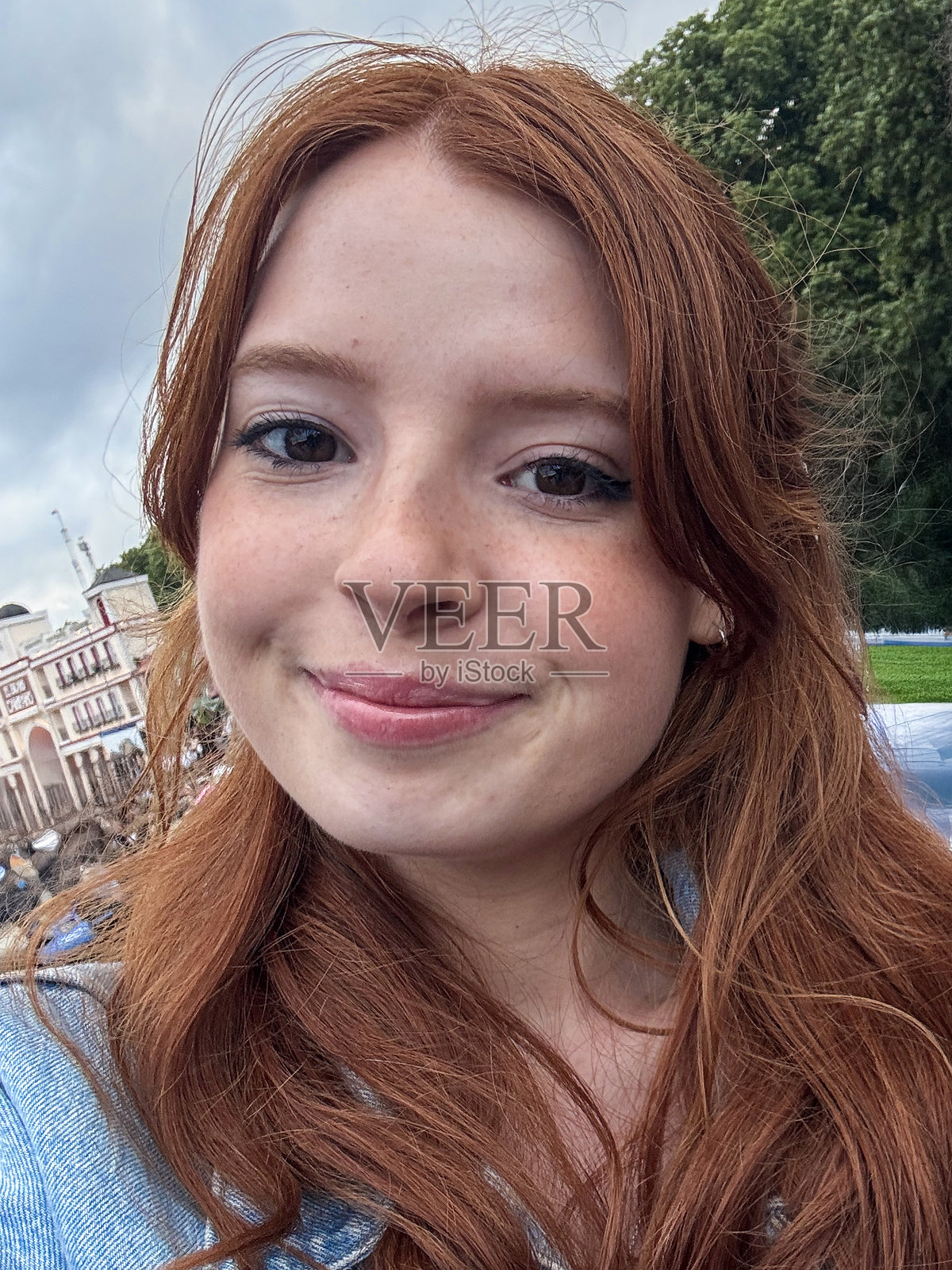 全景图像中,一位迷人的红发年轻女性在户外音乐会场地摆姿势,微笑着看向镜头,背景是灰色阴云的天空,焦点在前景。照片摄影图片