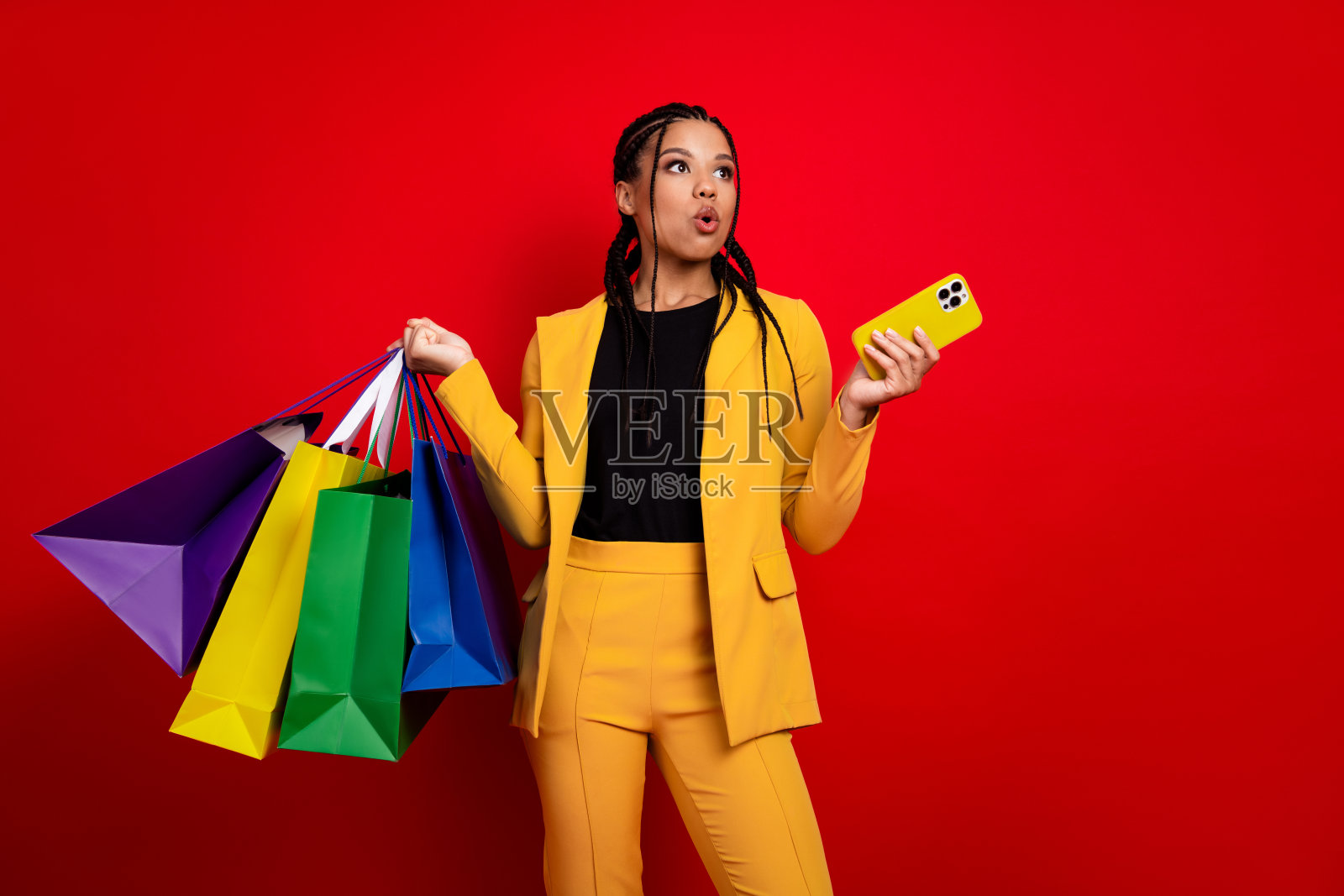 身穿黄色西装的优雅女性手持购物袋和手机,背景为鲜艳的红色,展示时尚与风格。照片摄影图片