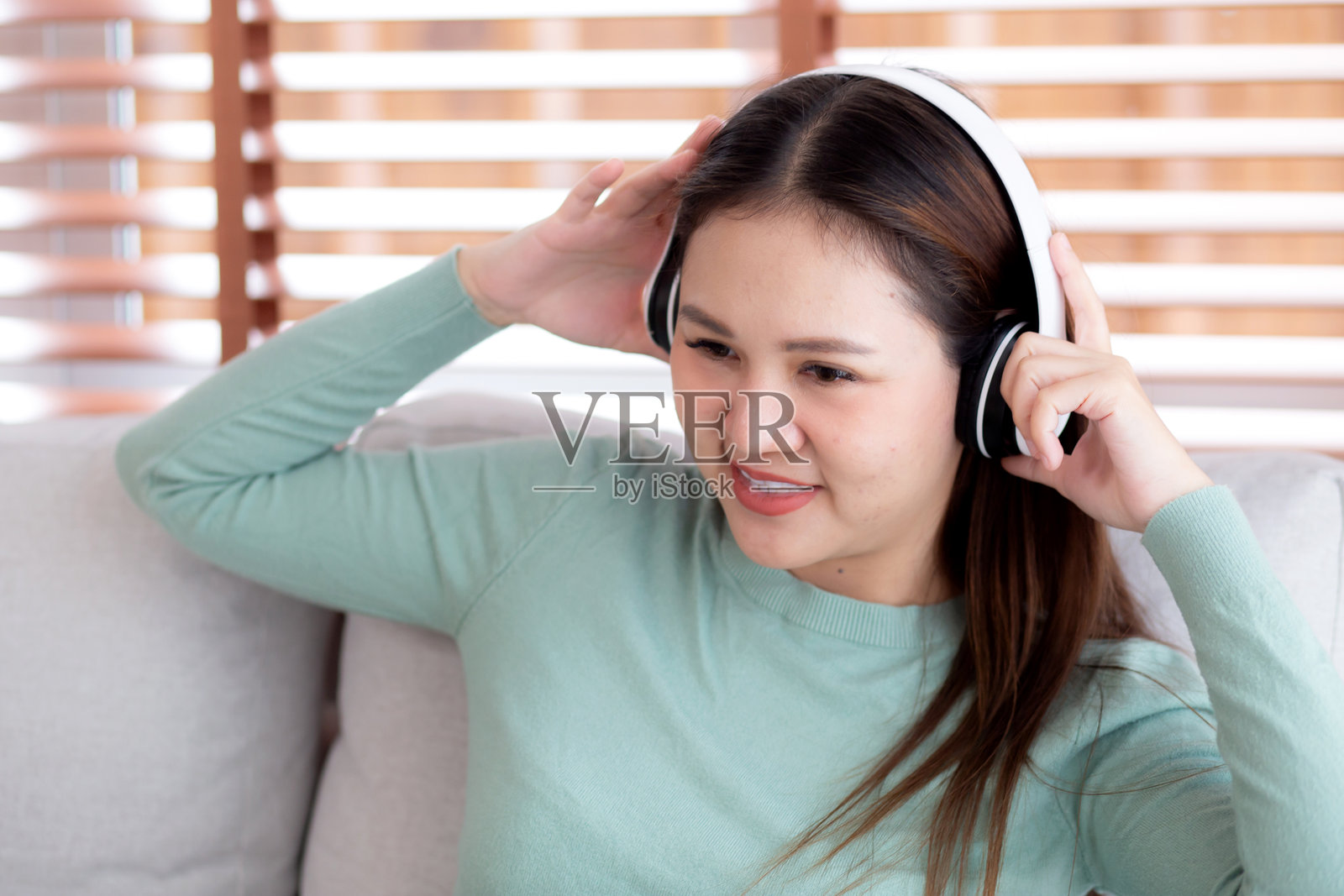 快乐的年轻亚洲女性坐在沙发上,戴着耳机听音乐,享受家庭客厅的乐趣,体现了女性的幸福与娱乐,生活方式的概念。照片摄影图片