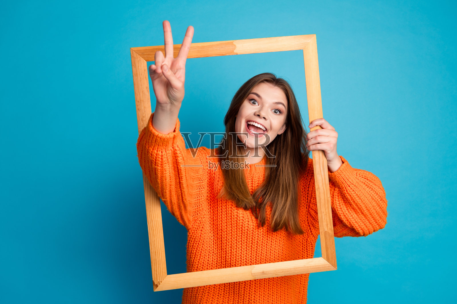 穿着橙色毛衣的年轻女性在蓝色背景下透过框架开心地微笑照片摄影图片