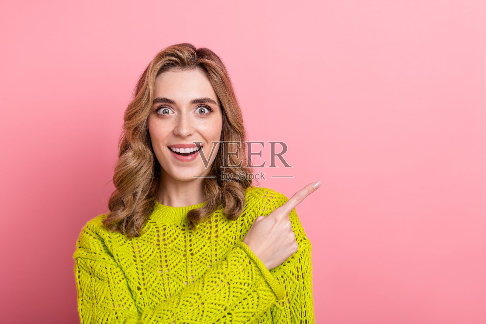 穿着黄色毛衣的快乐女性在鲜艳的粉色背景前兴奋地指着。照片摄影图片
