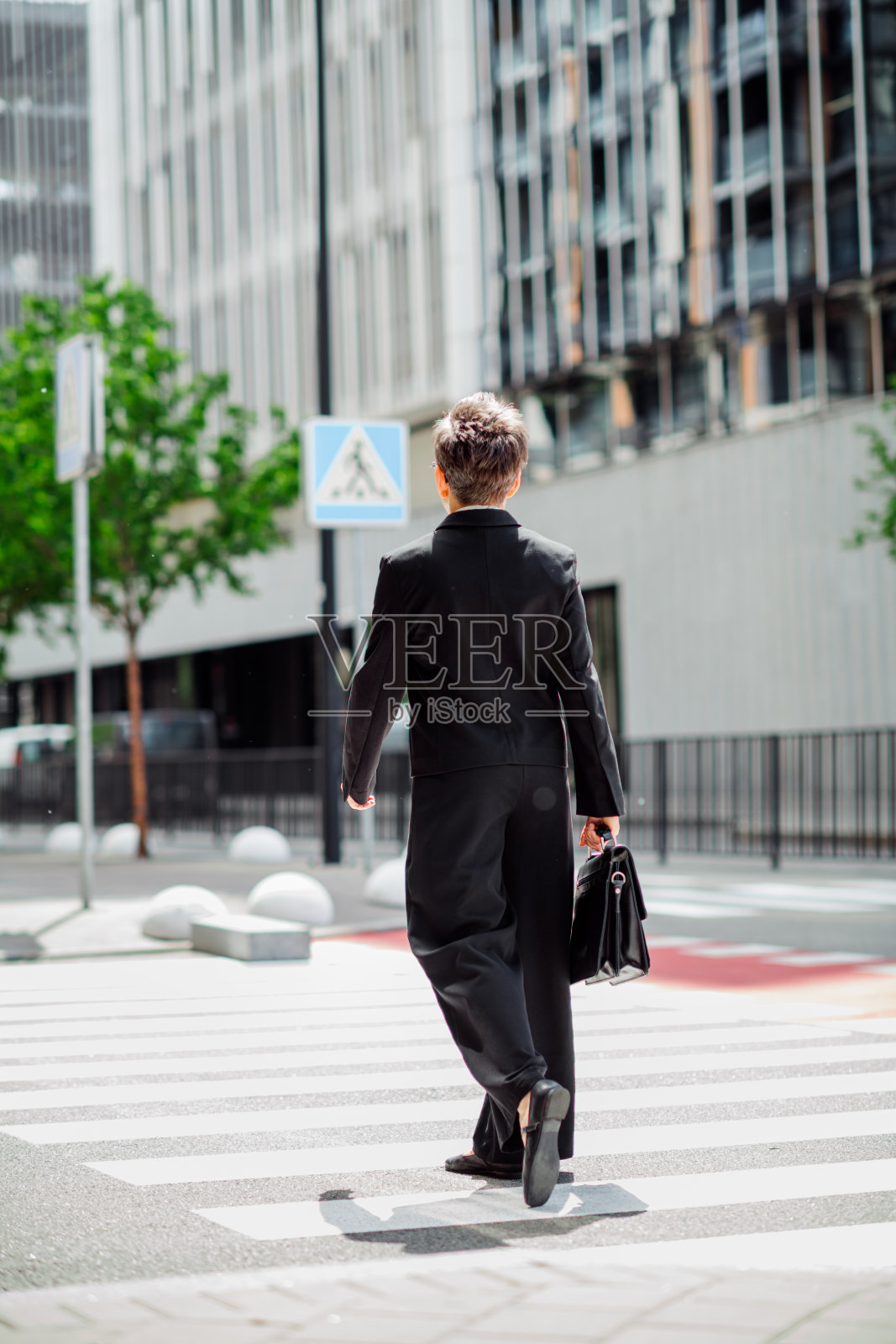 身穿正式服装的职业女性在现代城市风景中走过斑马线,手中提着一个包。照片摄影图片