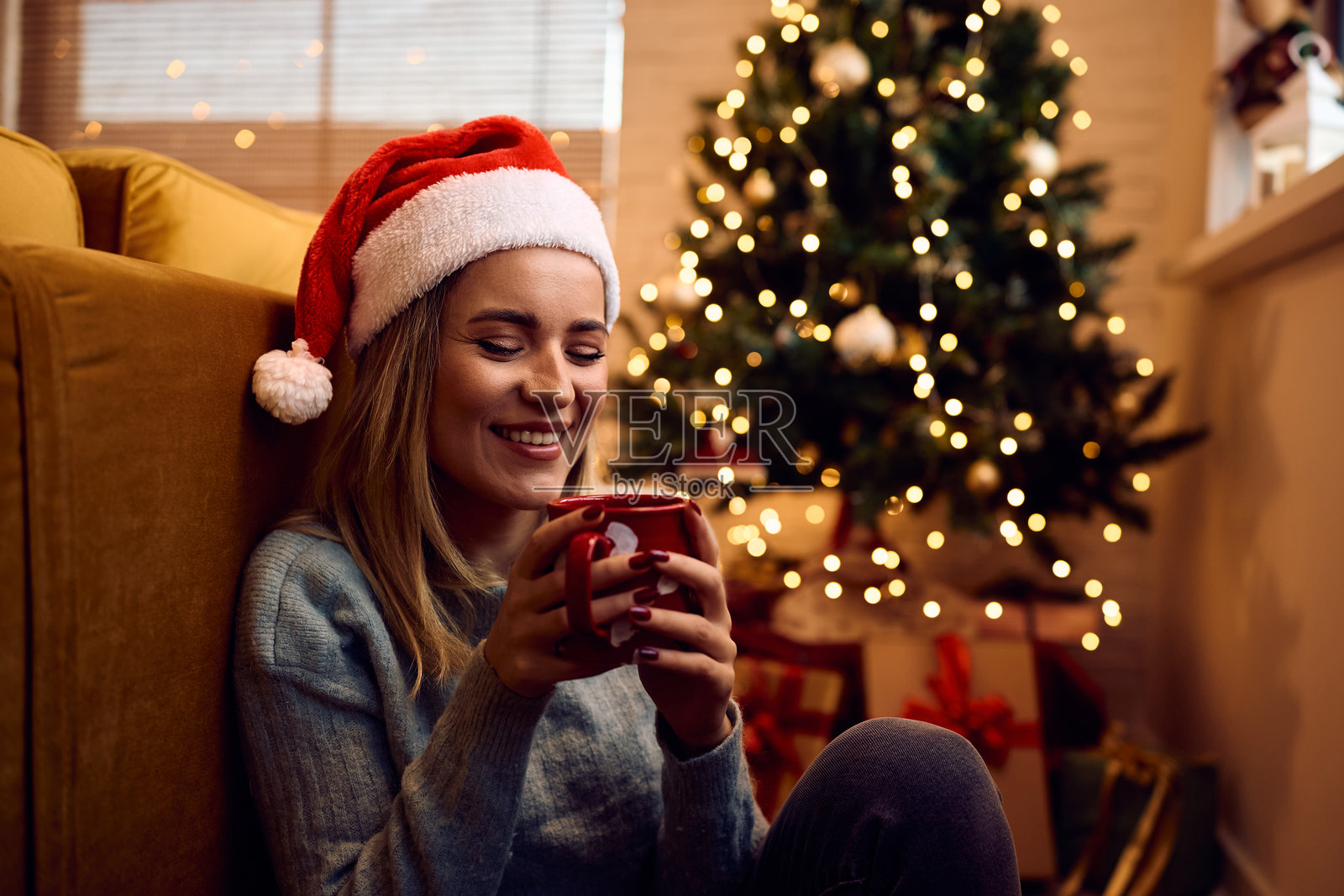 微笑的女人在家中圣诞树旁放松时喝茶。照片摄影图片