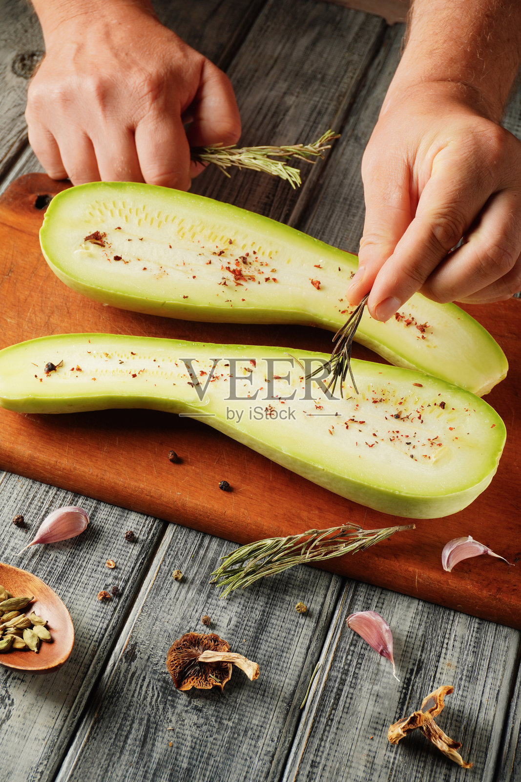 新鲜的绿色南瓜正在厨房中准备。手正在向切好的蔬菜上添加香草和香料,木砧板周围摆放着其他配料。照片摄影图片