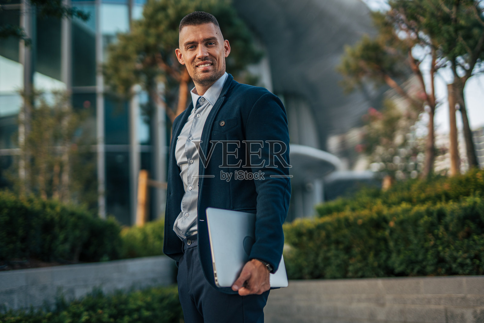 商人在办公大楼前微笑着拿着笔记本电脑对着镜头。照片摄影图片