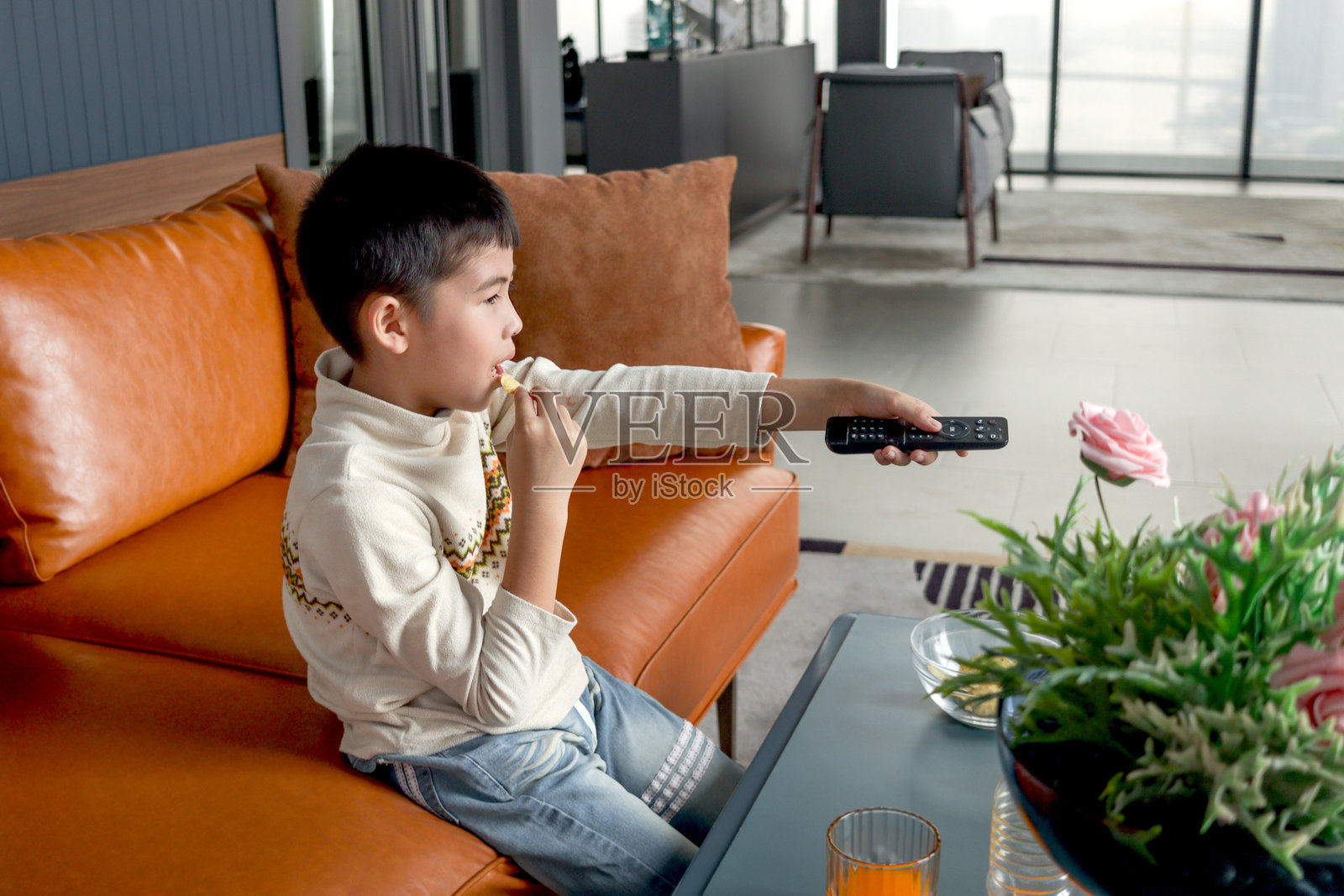 快乐的亚洲男孩手持遥控器换频道,坐在客厅沙发上看电视节目并吃零食。愉快的孩子在观看电影时度过美好时光。可爱的孩子在家中享受。照片摄影图片