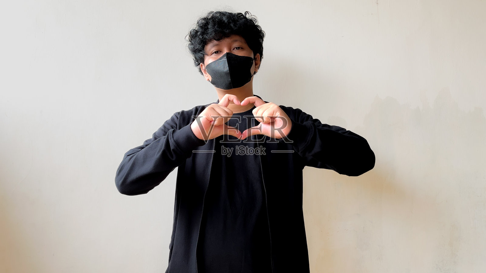 一名穿着黑色服装和面具的男子站在一面简单的墙前,用双手拇指和食指相连做出心形手势。照片摄影图片