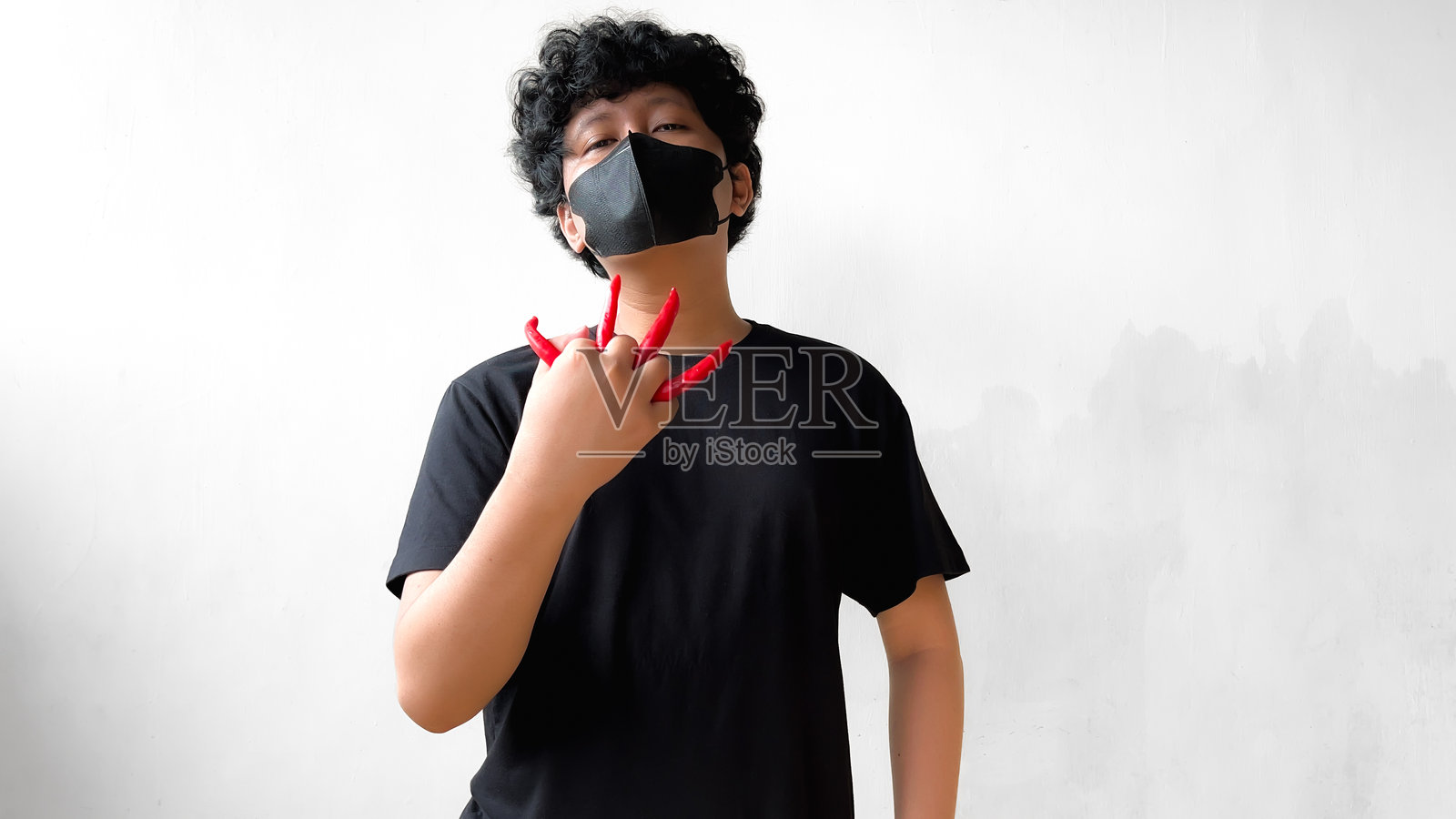一名男子戴着黑色口罩和黑色T恤,手指上夹着红辣椒,展现出一种顽皮独特的手势。照片摄影图片