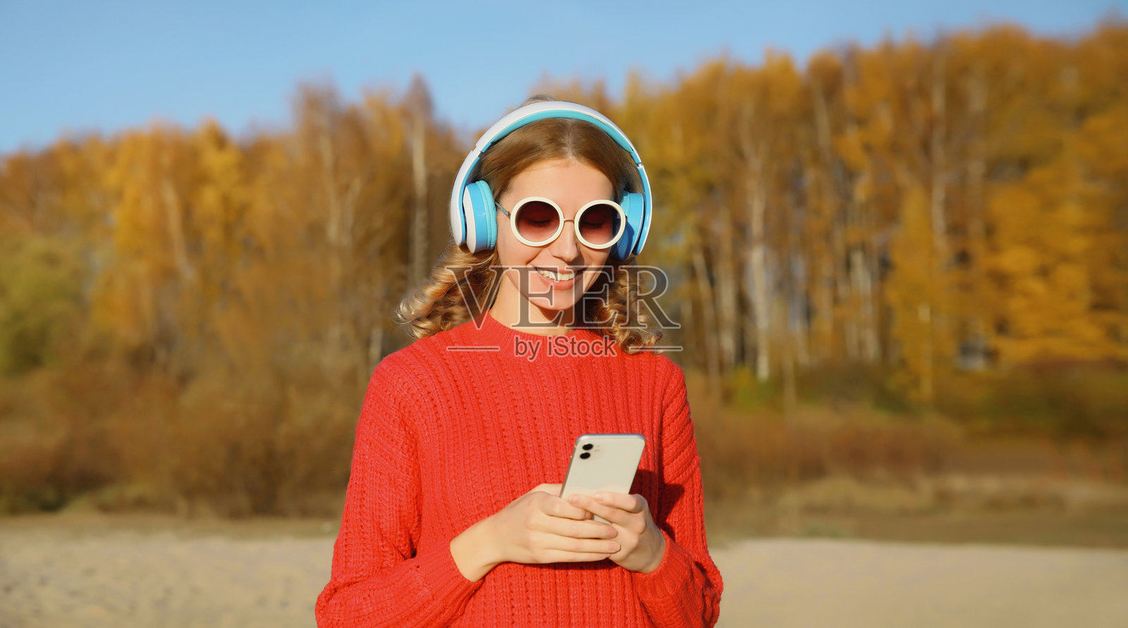 快乐的年轻女性在秋季公园户外戴着耳机享受音乐,手里拿着手机。照片摄影图片