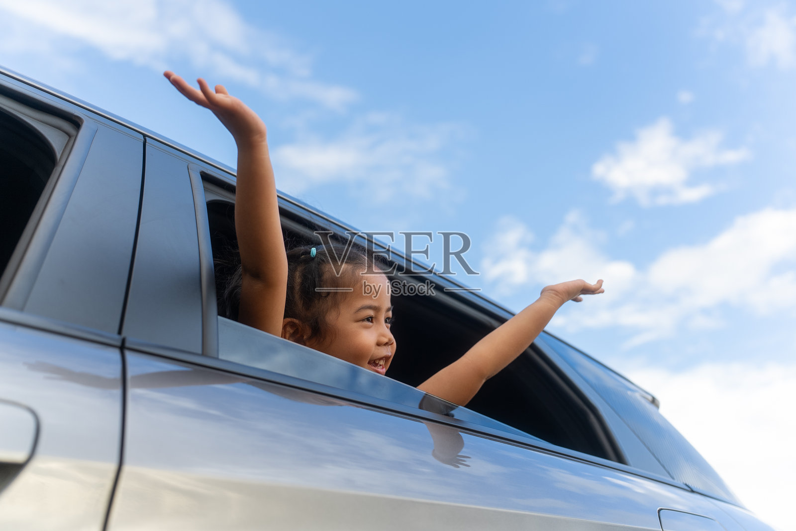 快乐的卷发女孩在夏日家庭公路旅行中,兴奋地把手伸出开着的车窗,侧面视角展示了她与蓝天背景下的激动旅程。照片摄影图片