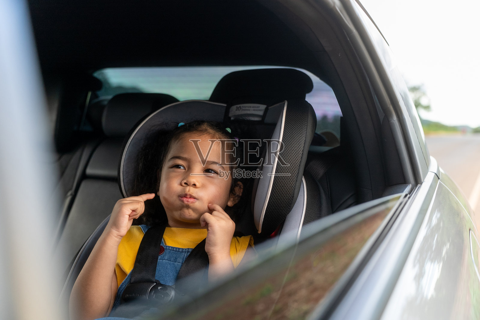 快乐的卷发小女孩坐在后座安全带上,笑着、开心地望着开着的车窗,享受着家庭夏季公路旅行的时光。照片摄影图片