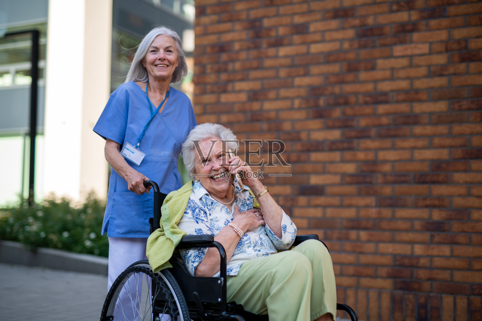 护理人员推着一位正在打电话的老年女性坐在轮椅上。照片摄影图片