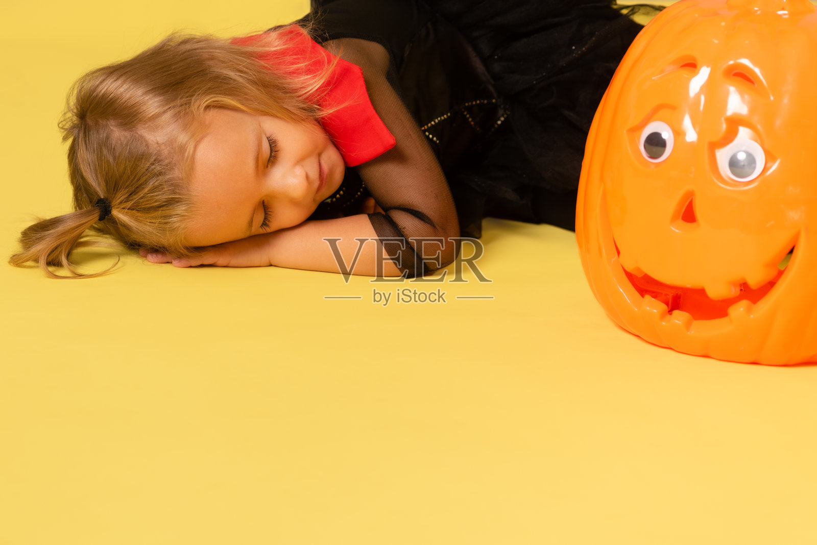 穿着万圣节服装的女孩躺在黄色地板上,身边有一个塑料南瓜,横向留白空间照片摄影图片